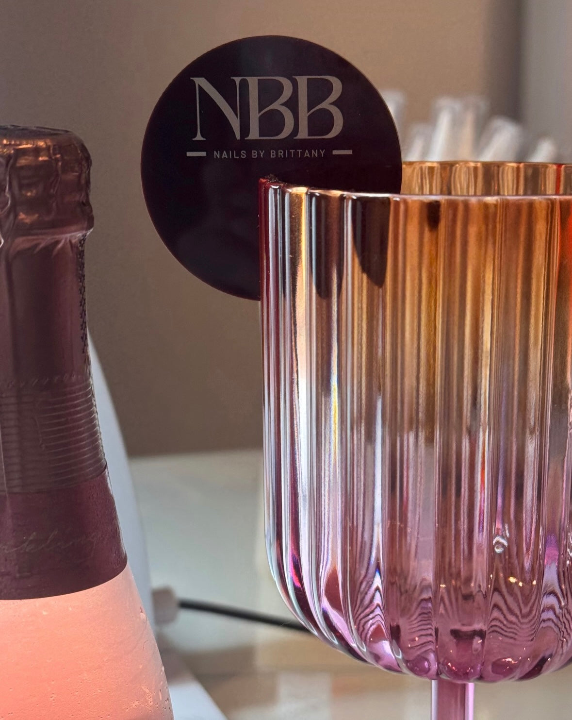 Custom round acrylic drink tag with NBB Nails by Brittany logo, displayed on the rim of an iridescent pink glass beside a champagne bottle.