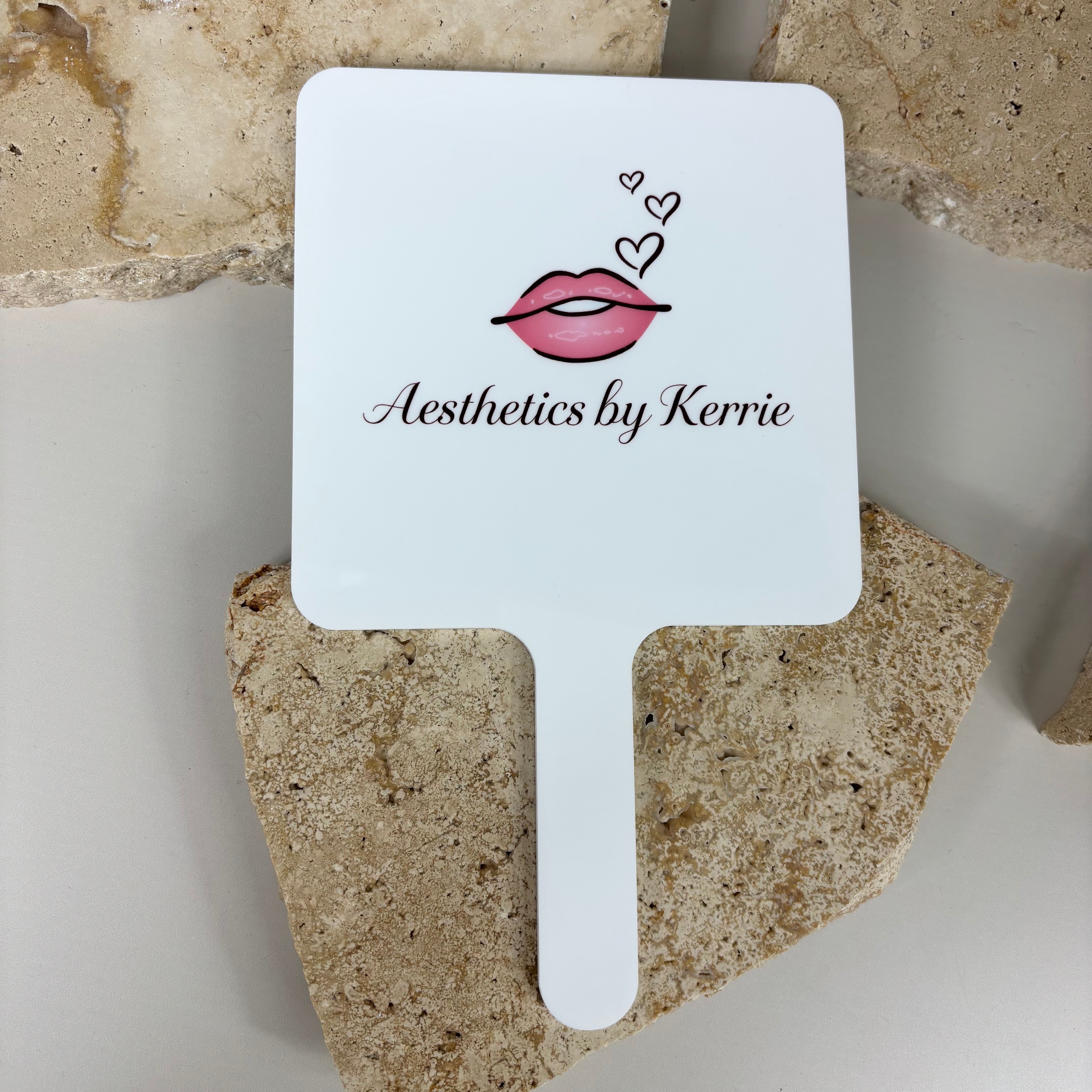 Custom white handheld salon mirror with “Aesthetics by Kerrie” branding, featuring a pink lips and hearts graphic, displayed on a neutral stone background.