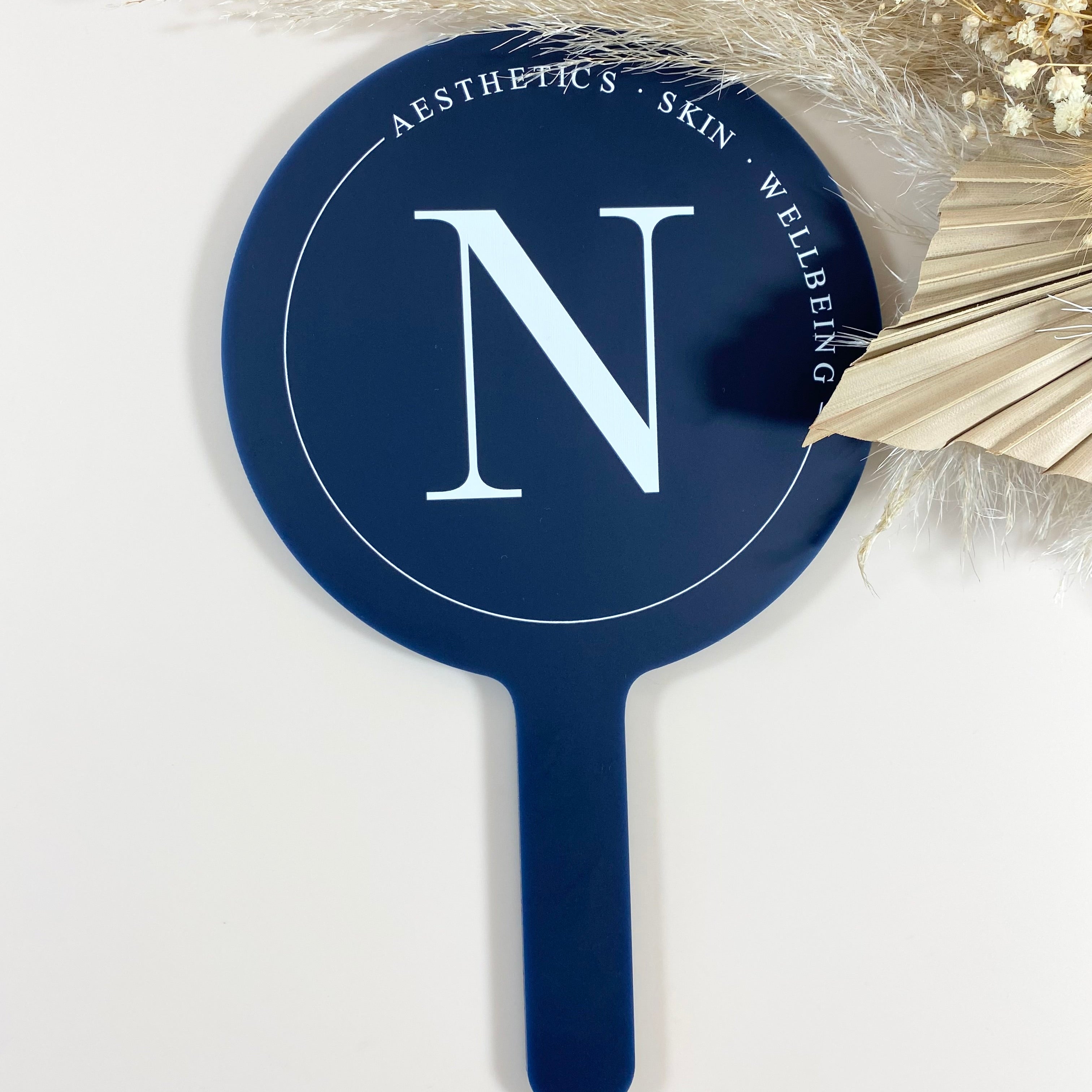 A round blue handheld mirror with customizable branding space, displayed with a feather decoration in the background.