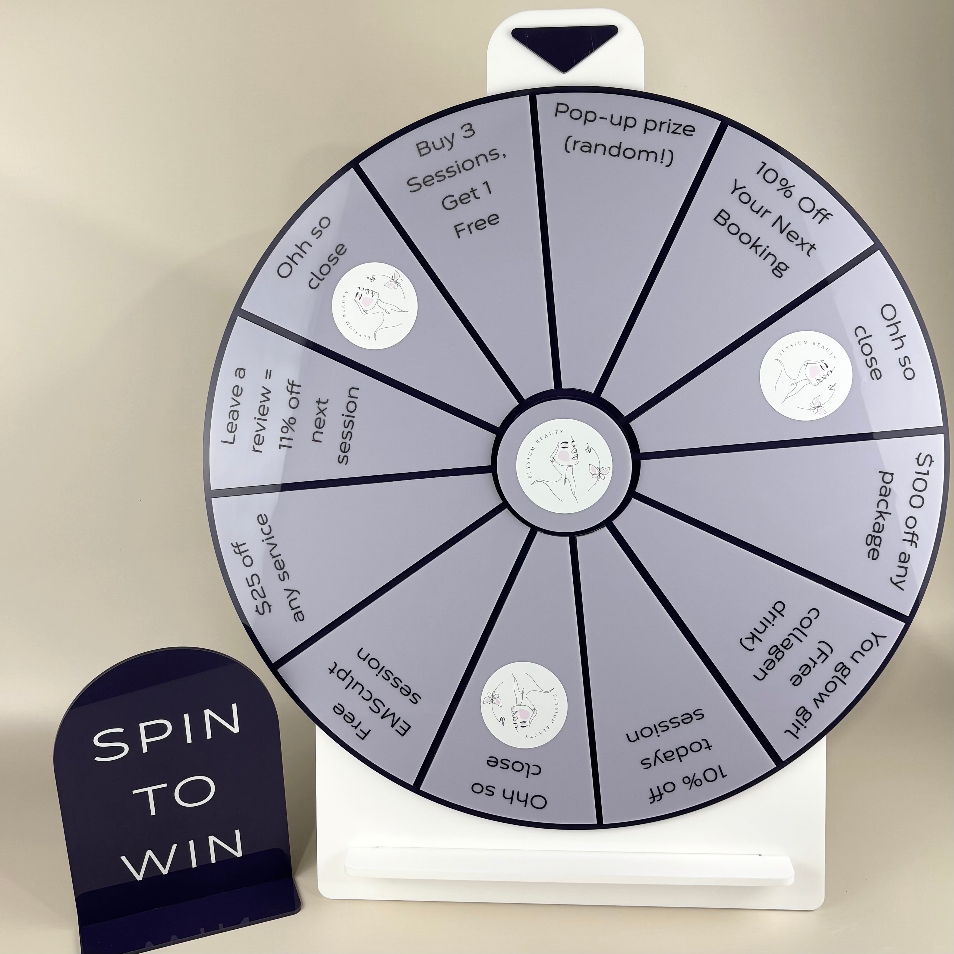 Custom acrylic “Spin to Win” prize wheel with grey and black segments featuring various offers and discounts for a beauty business, displayed beside a matching navy acrylic sign reading “Spin to Win.”