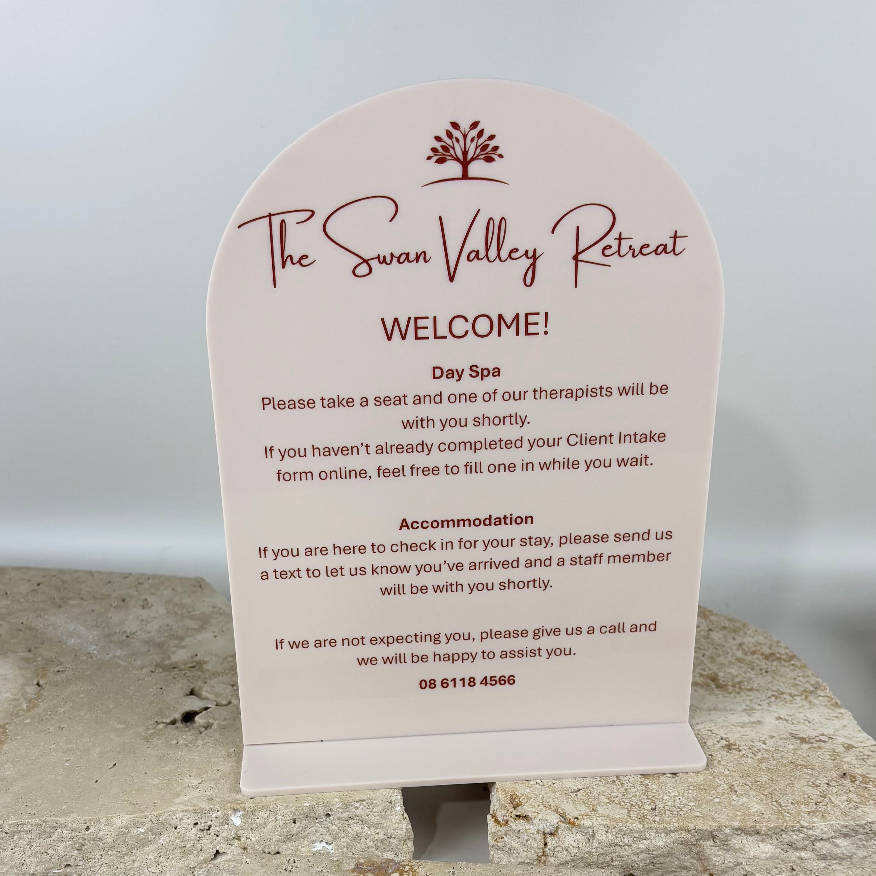 Frosted acrylic welcome sign for The Swan Valley Retreat day spa, featuring curved arch design with printed logo, guest check-in instructions and accommodation information, displayed on a reception counter in a luxury wellness spa setting.