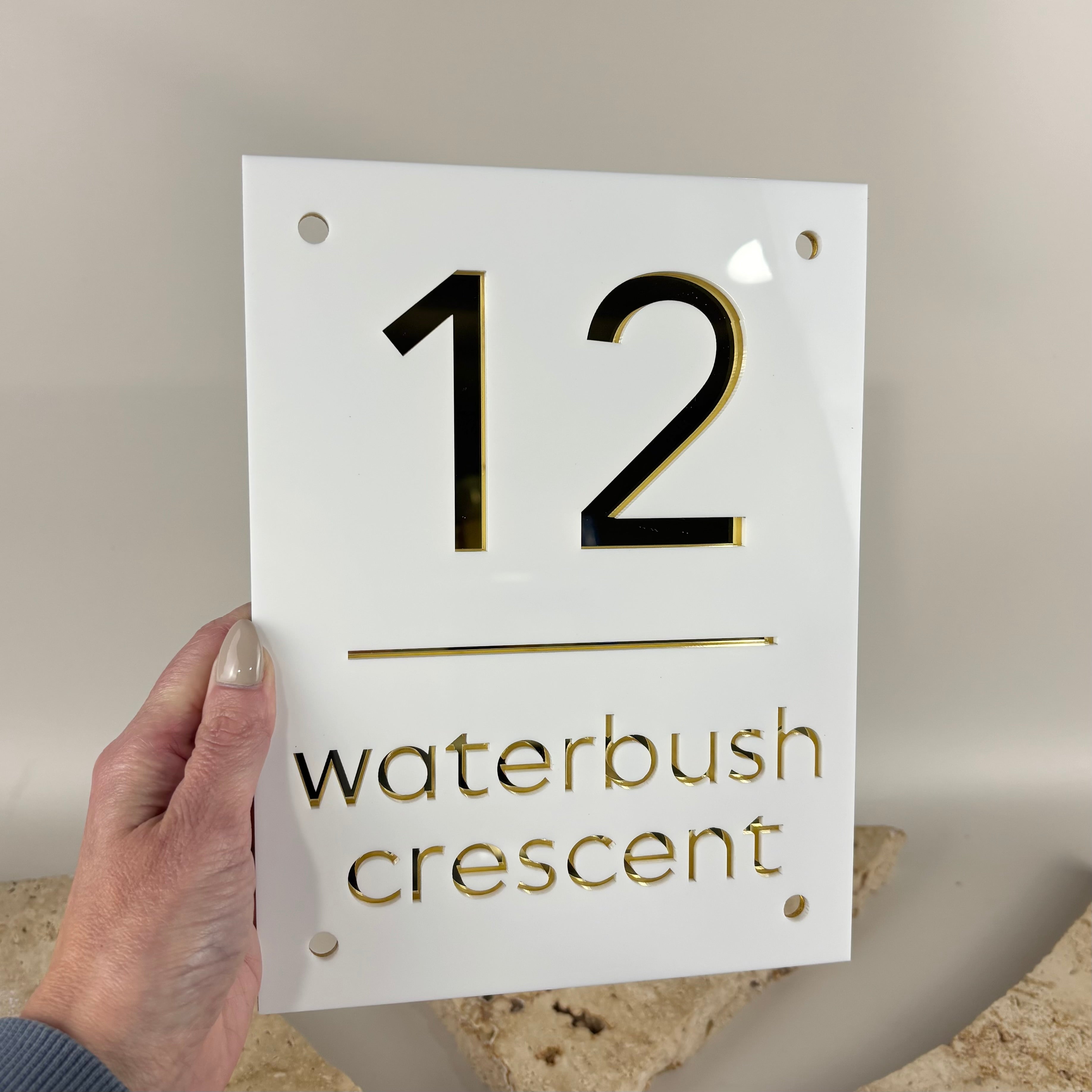 Modern white acrylic house number sign featuring laser-cut black and gold mirrored acrylic number 12 with Waterbush Crescent street name, mounted with silver standoff fixings. Custom contemporary address plaque for home entrance, front facade or letterbox.