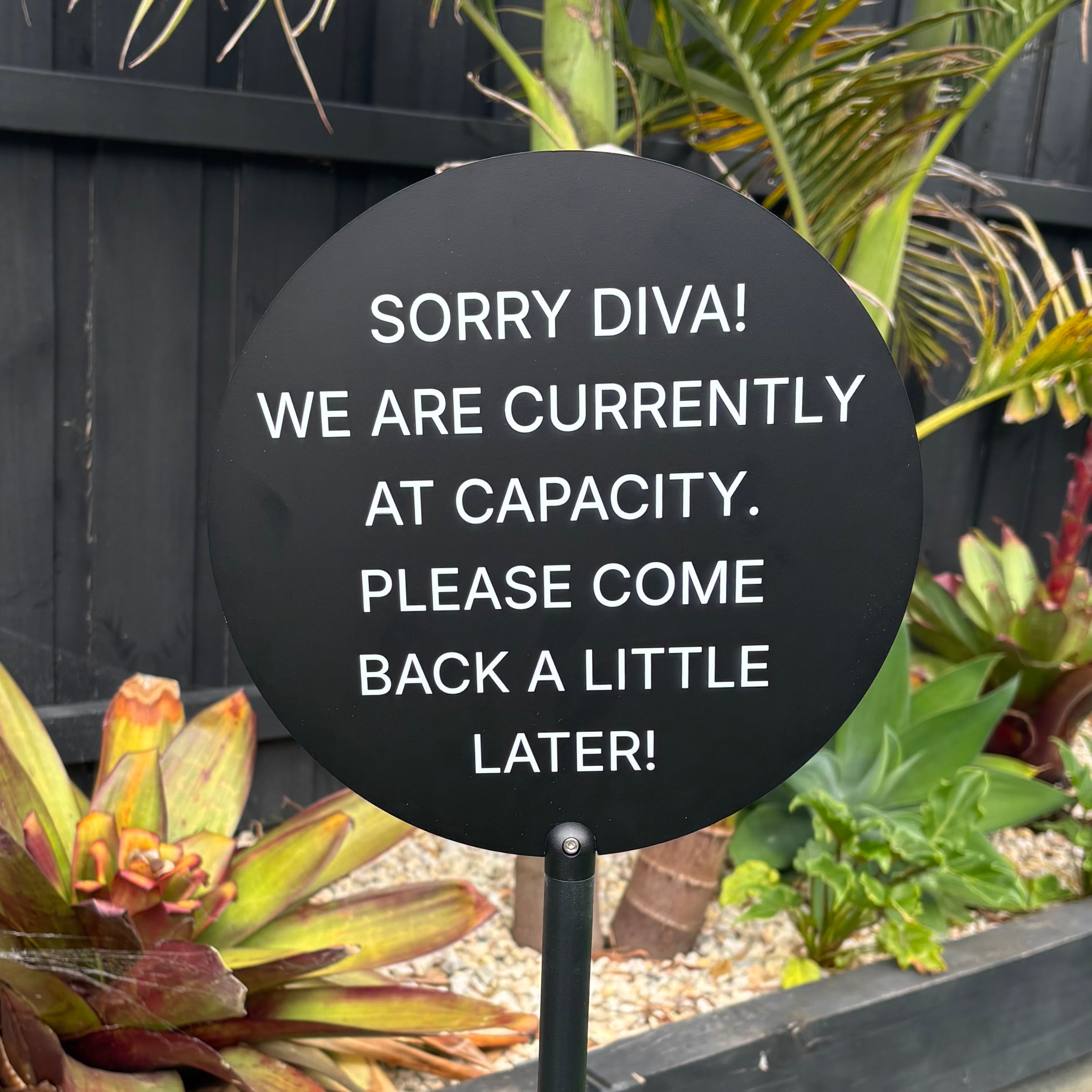 Black round metal floor sign with custom capacity message, freestanding outdoor business signage for crowd control and entry management, durable powder coated stand for retail and salon use.