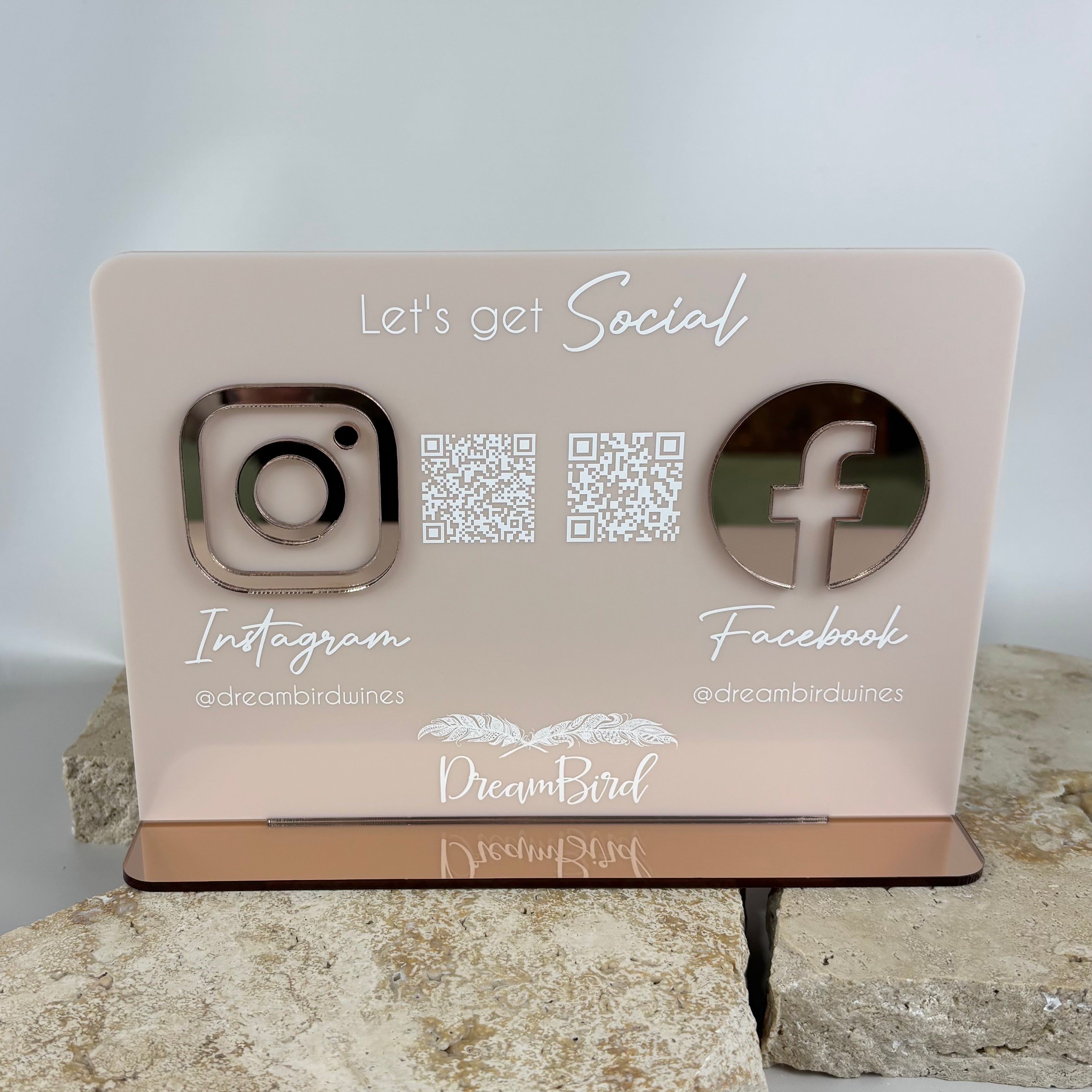 Blush pink acrylic social media display sign with rose gold Instagram and Facebook icons, QR codes, and white text reading “Let’s get Social” and “DreamBird @dreambirdwines,” placed on stone slabs with a glossy rose gold base.