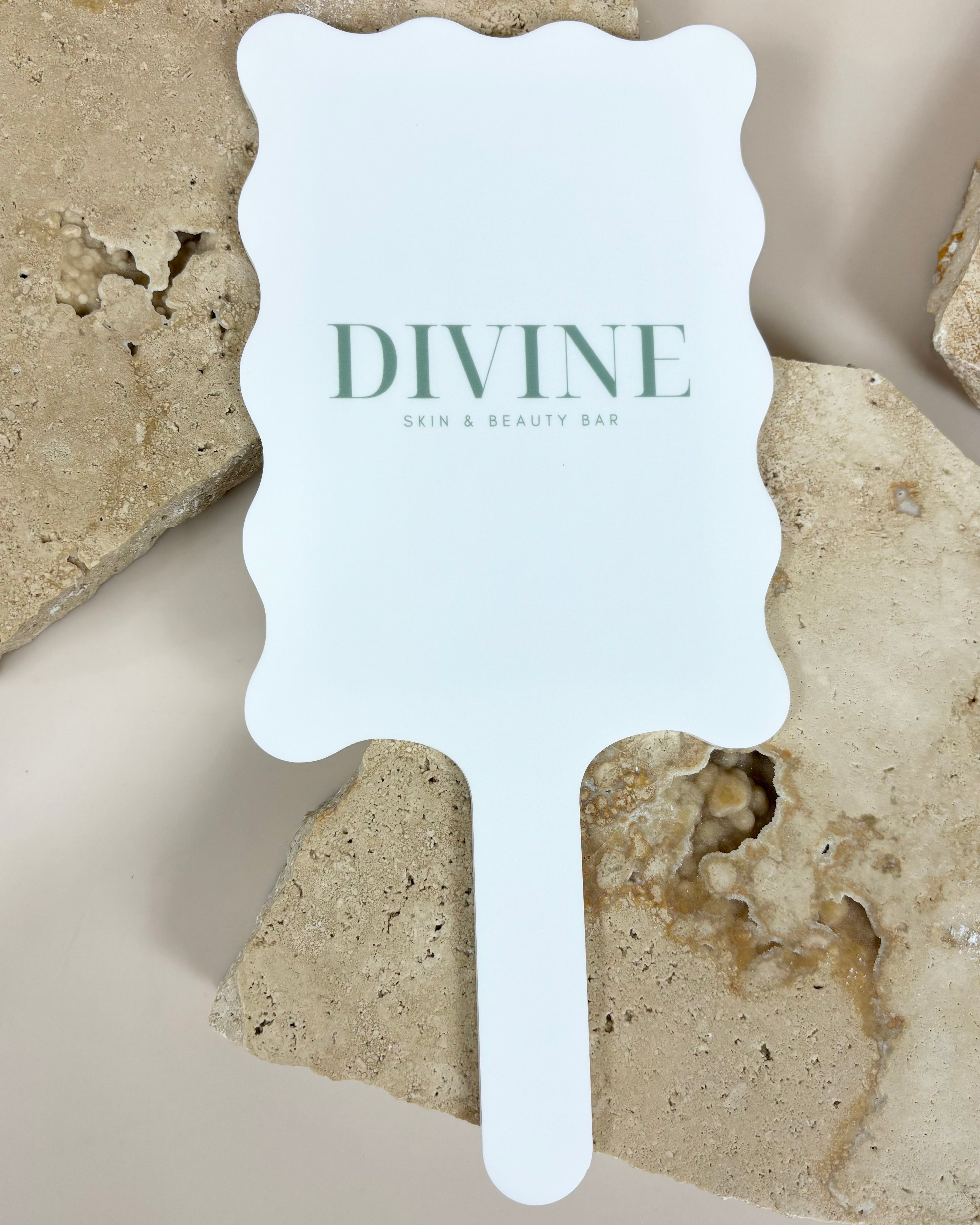 Branded white wavy handheld mirror with Divine logo displayed on stone background