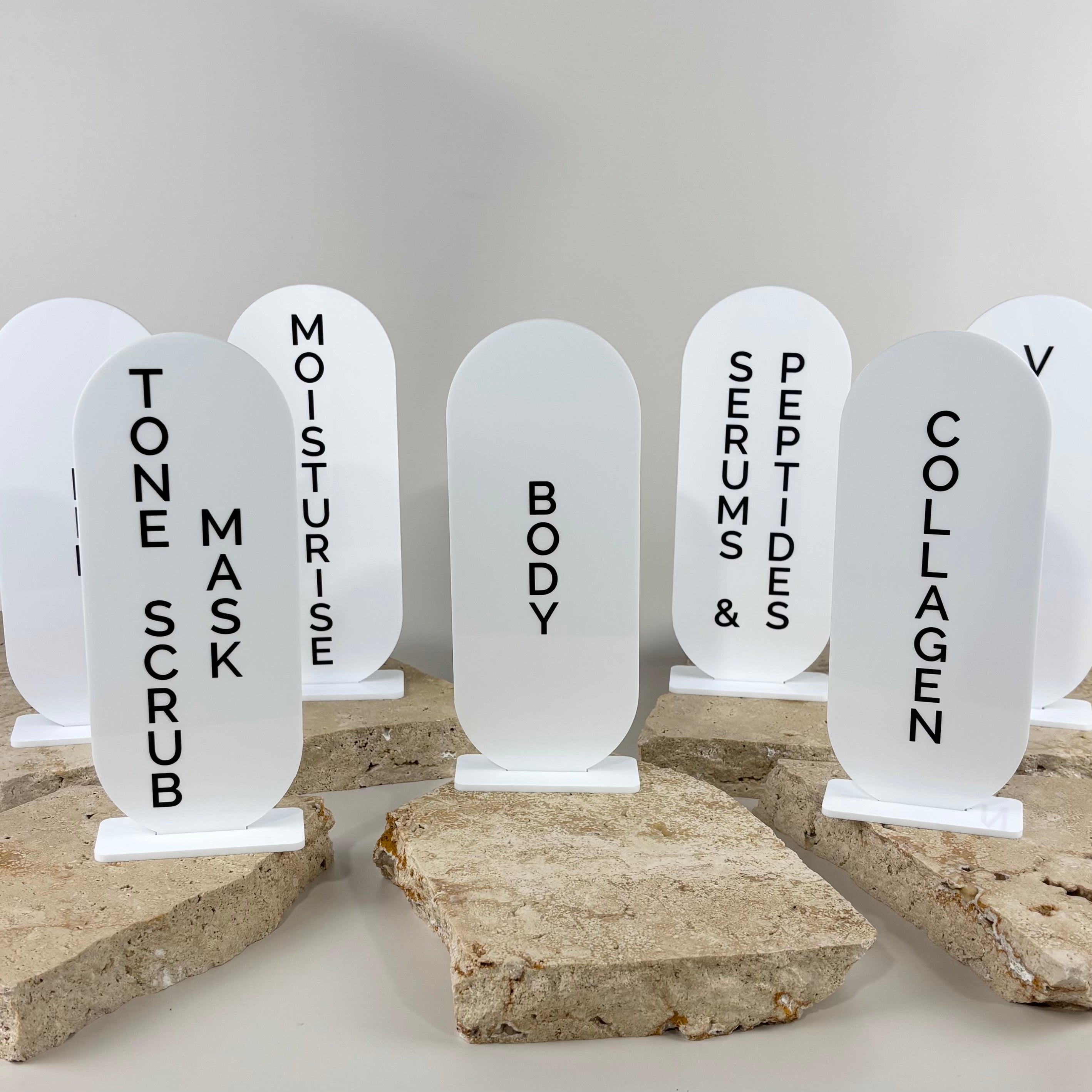 Minimal white acrylic product category display signs for serums, collagen, moisturiser and skincare products in a luxury clinic display setup