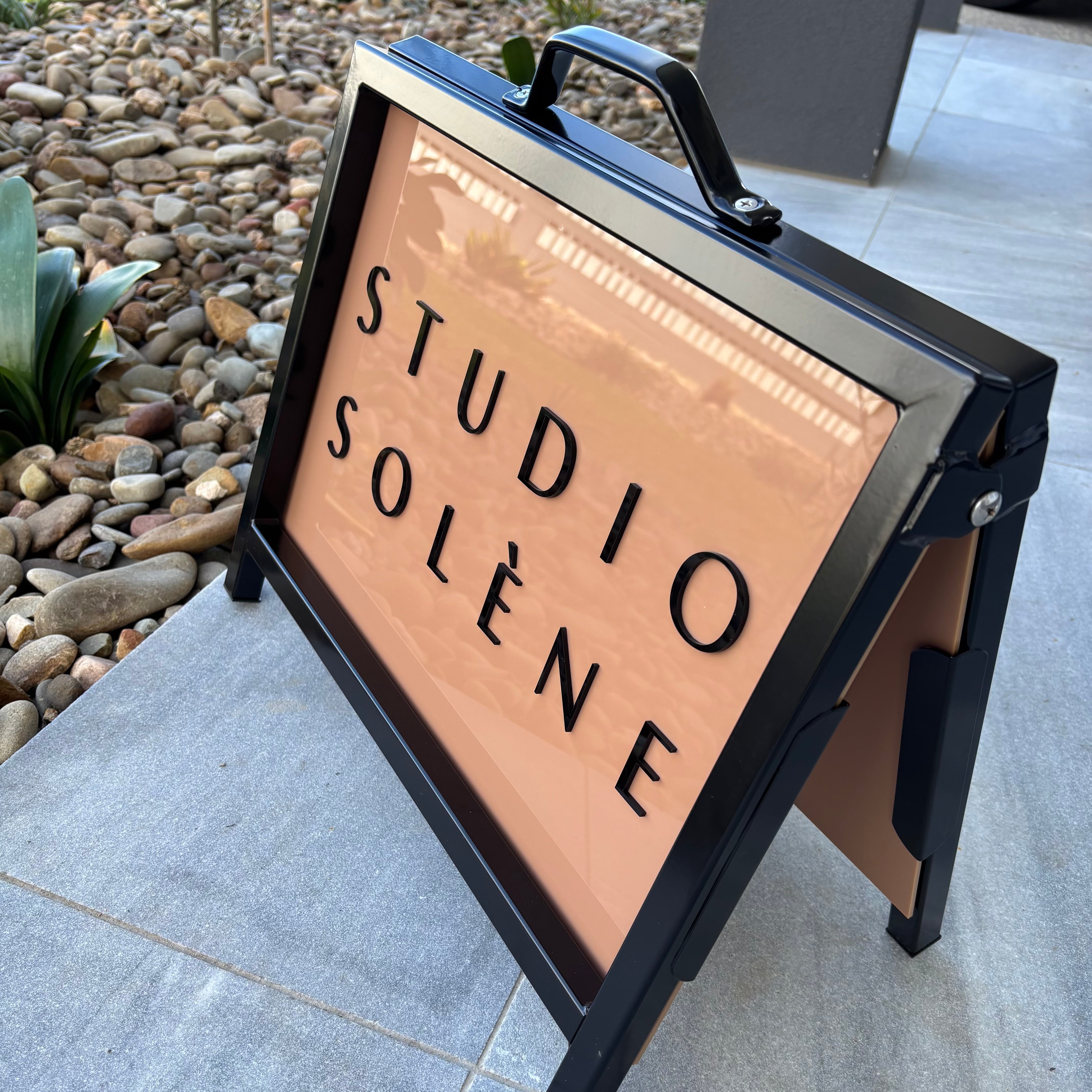 Custom portable A-frame sign for “Studio Solène” featuring a peach acrylic panel with black 3D lettering in a black metal frame, positioned on a tiled entryway with decorative garden stones.