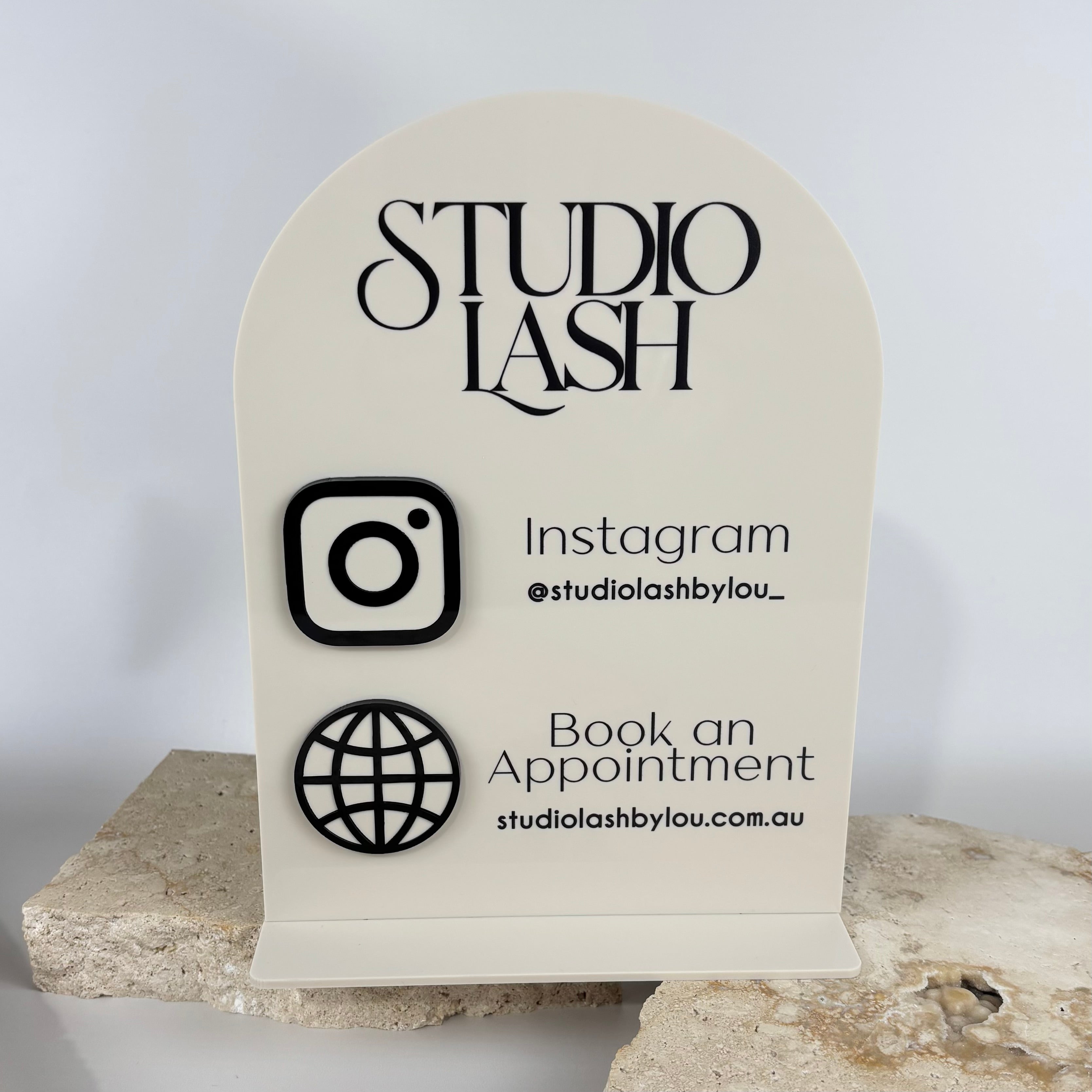 NFC acrylic social media display sign for Studio Lash, featuring black raised Instagram and website icons with text “@studiolashbylou_” and “Book an Appointment studiolashbylou.com.au,” displayed on neutral stone slabs.