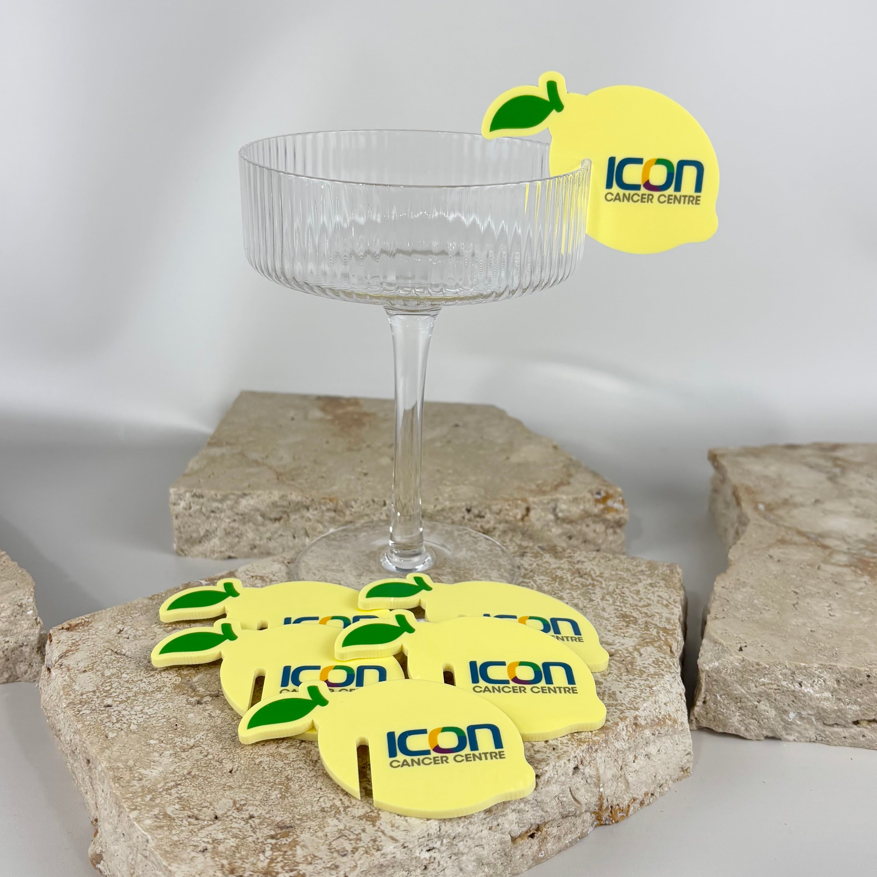 Lemon-shaped acrylic drink tags featuring the “ICON Cancer Centre” logo, displayed on and around a clear ribbed glass coupe placed on textured stone slabs.
