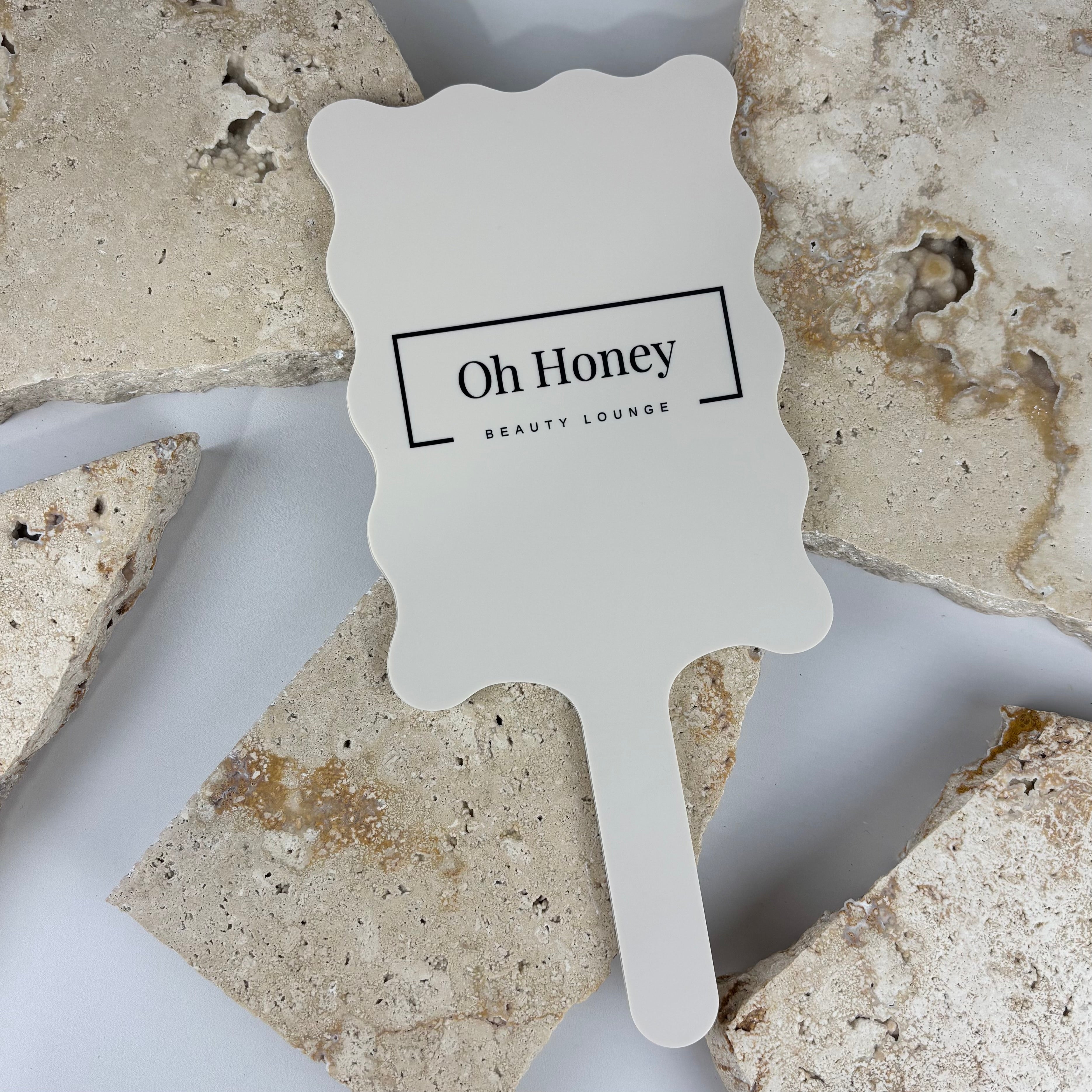 White acrylic wavy-edge handheld mirror featuring black logo text reading Oh Honey Beauty Lounge, styled flat lay on neutral stone tiles