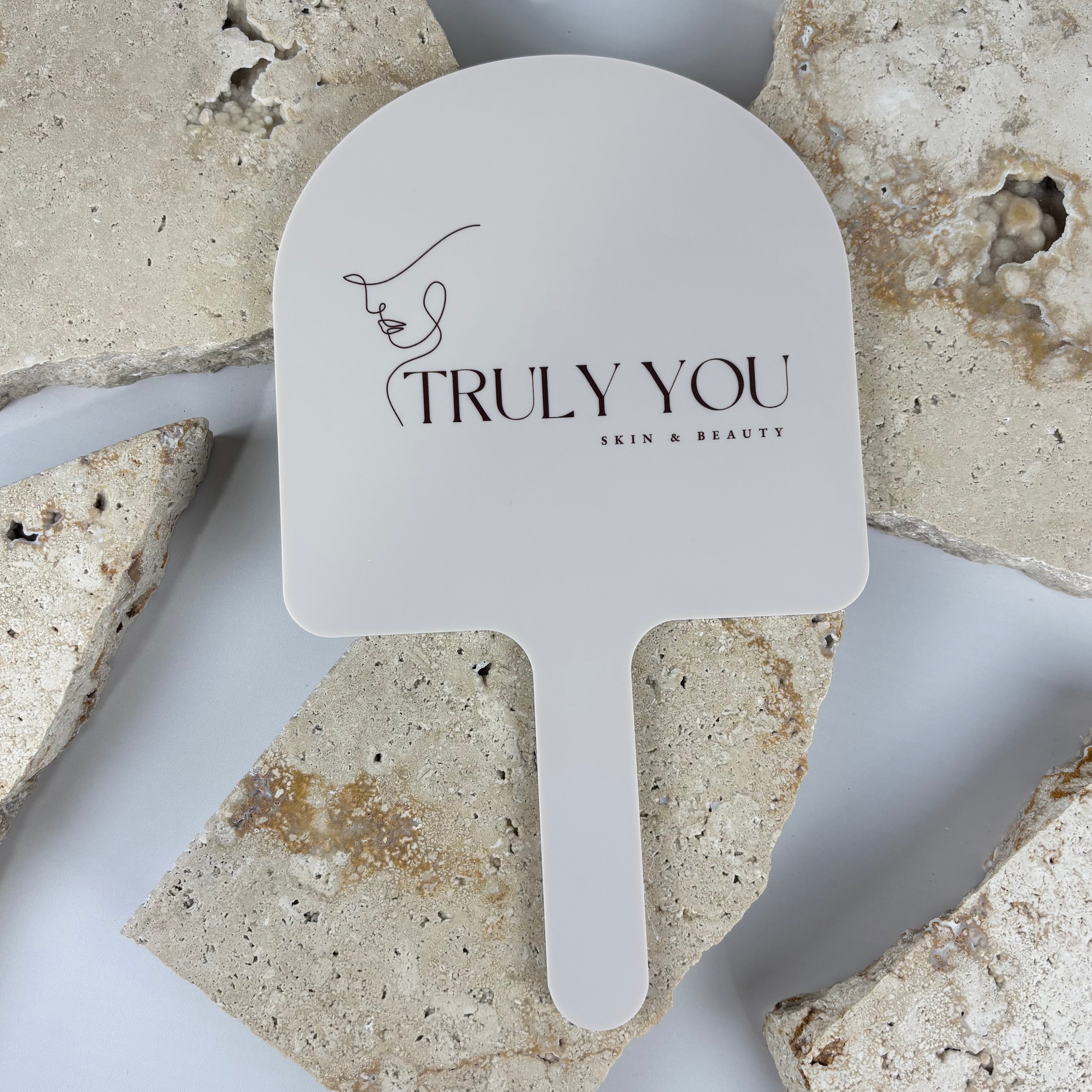 White acrylic arch-shaped handheld mirror with minimalist face line logo and text reading Truly You Skin & Beauty, photographed on a textured stone backdrop