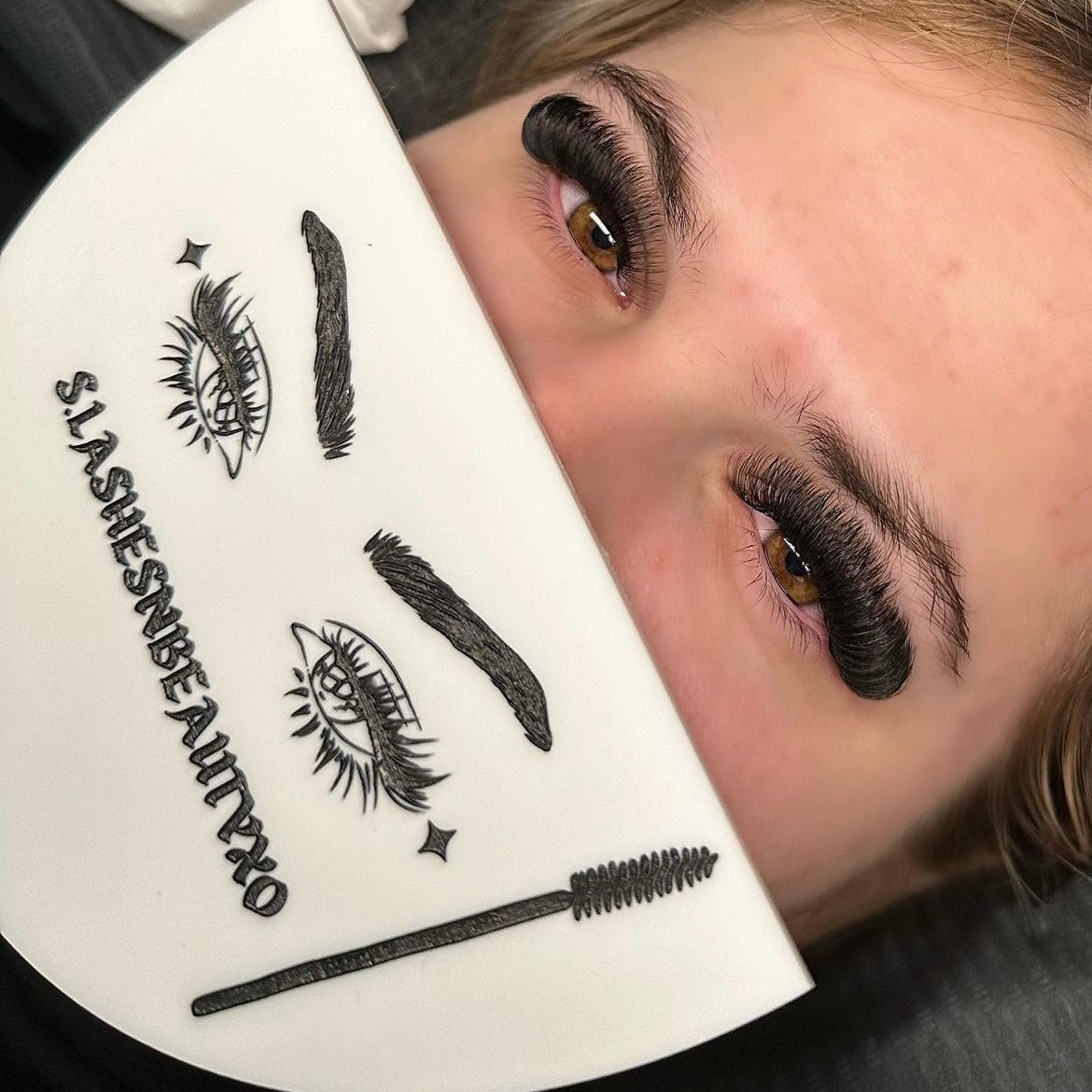 Lash artist holding a white branded face shield with eyelash graphics