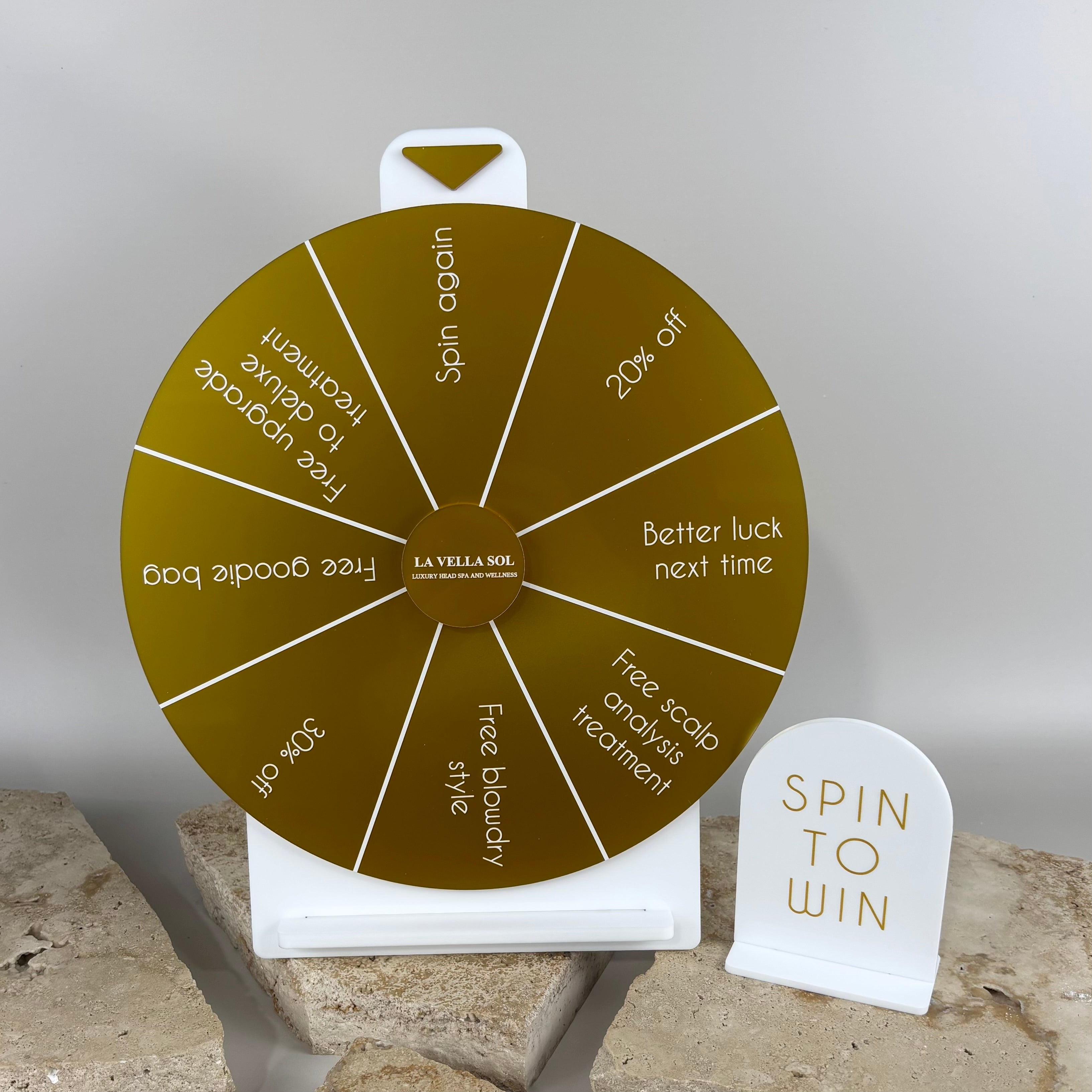 Gold acrylic spin to win prize wheel with white engraved sections displaying salon rewards such as discounts, free treatments and giveaways, paired with a matching white acrylic “Spin to Win” sign, displayed on stone blocks for La Vella Sol hair and wellness salon.