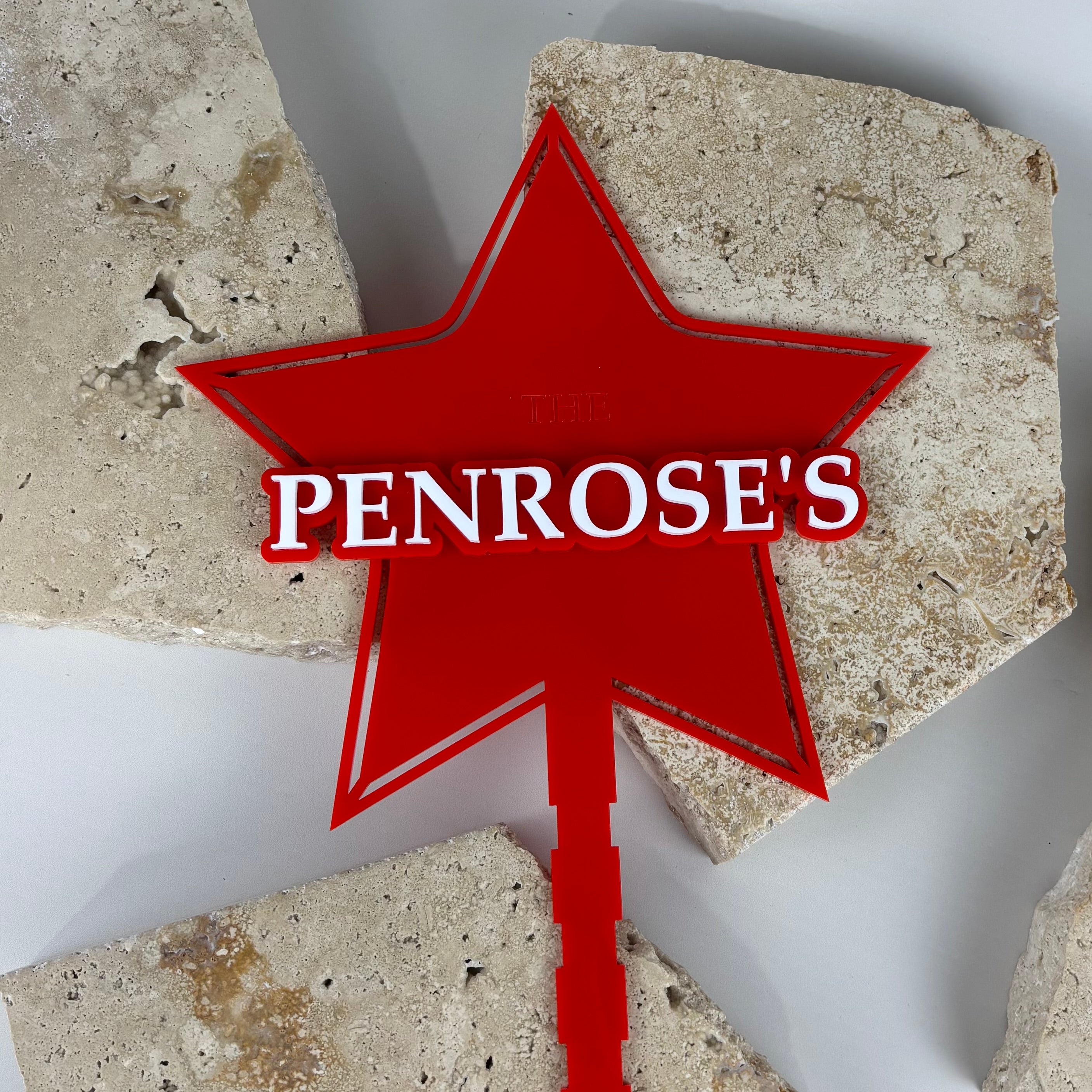 Custom red acrylic star-shaped Christmas tree topper with 3D white surname text reading “Penrose’s,” personalised holiday decor displayed on natural stone background.