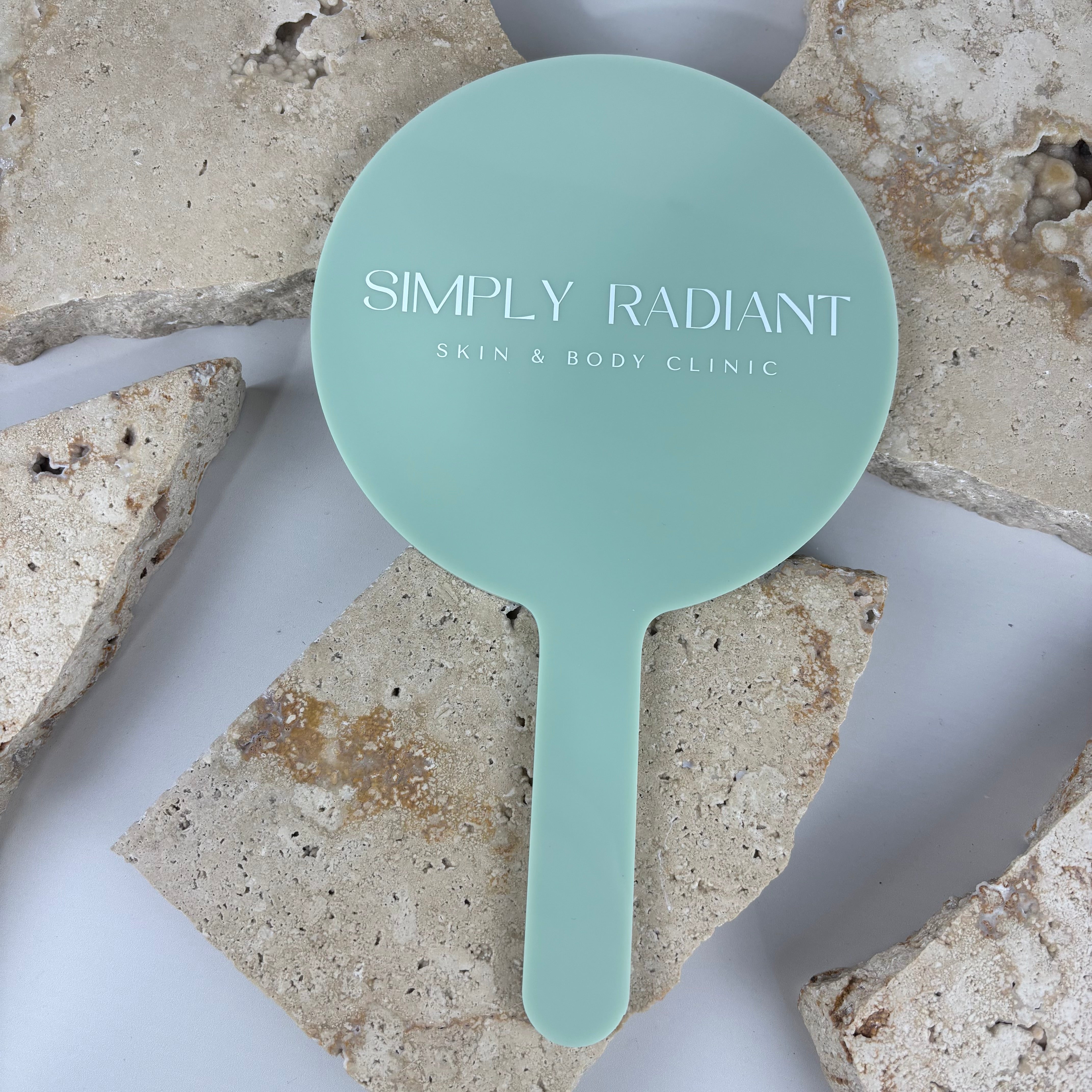 Mint green acrylic handheld mirror with white logo text reading Simply Radiant Skin & Body Clinic, photographed on a neutral stone background