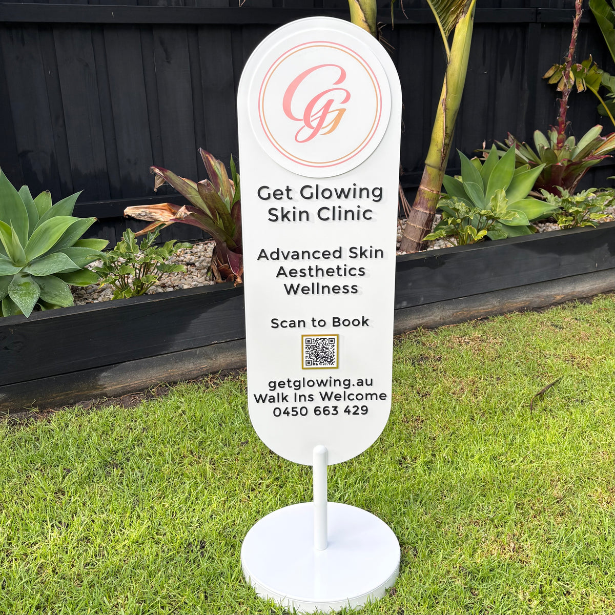 Custom oval freestanding business sign with UV printed logo, raised 3D acrylic lettering and QR code for Get Glowing Skin Clinic, featuring a premium white acrylic display stand for beauty salons, cosmetic clinics, medical aesthetics and retail storefront branding.