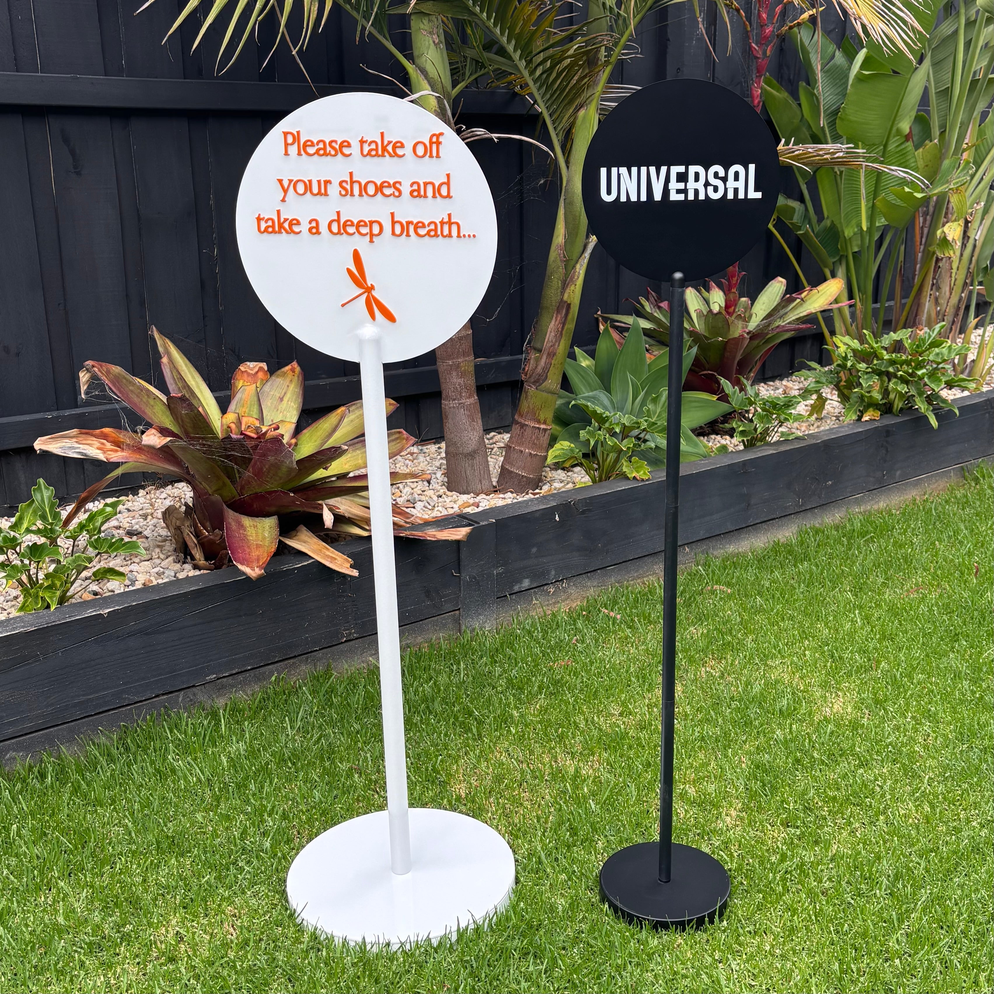 Set of freestanding round acrylic signs on metal stands, white sign with 3D orange lettering and black sign with bold “Universal” text, modern outdoor business signage, custom acrylic display signs for events or storefronts
