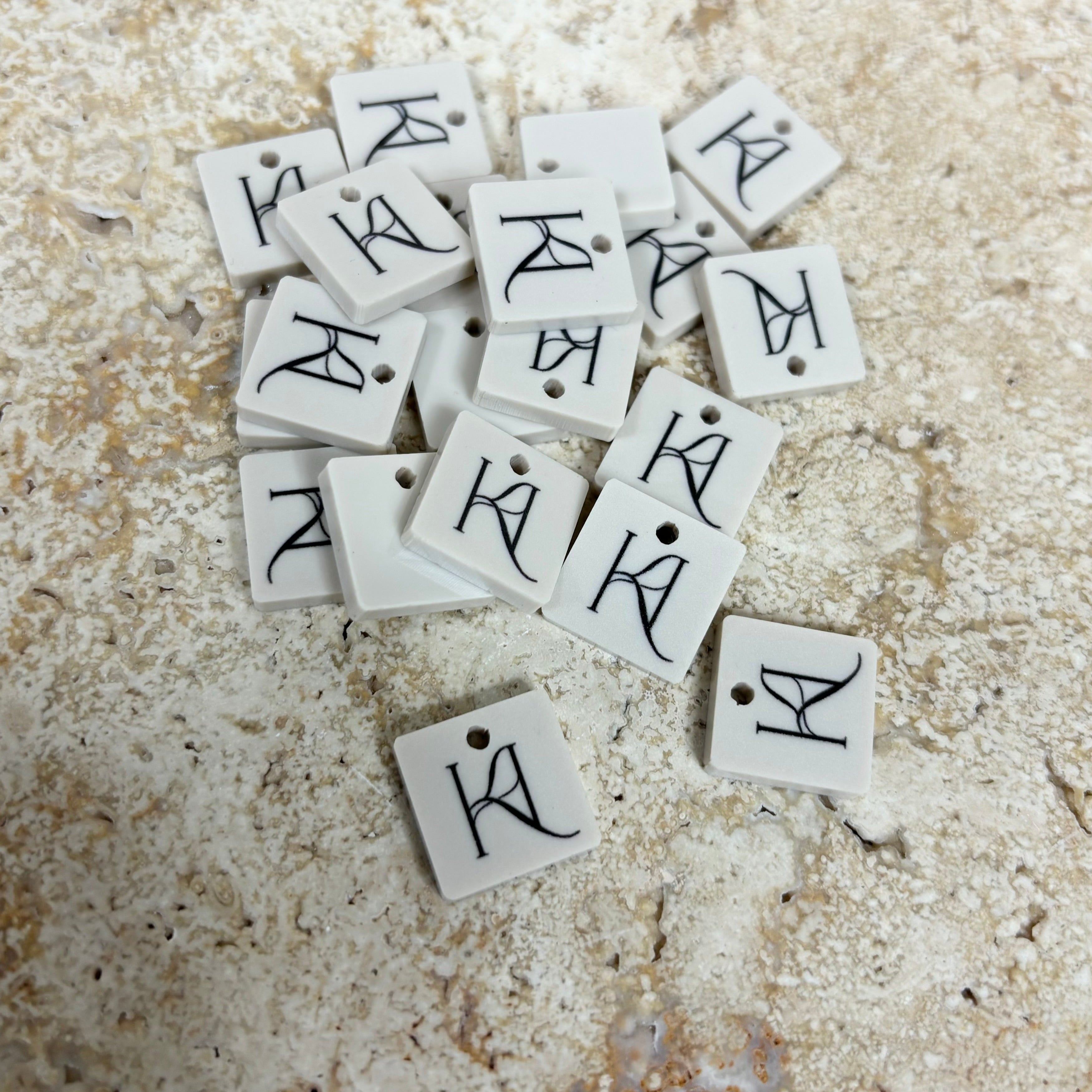 Pile of square beige acrylic product tags with engraved initials for crafters