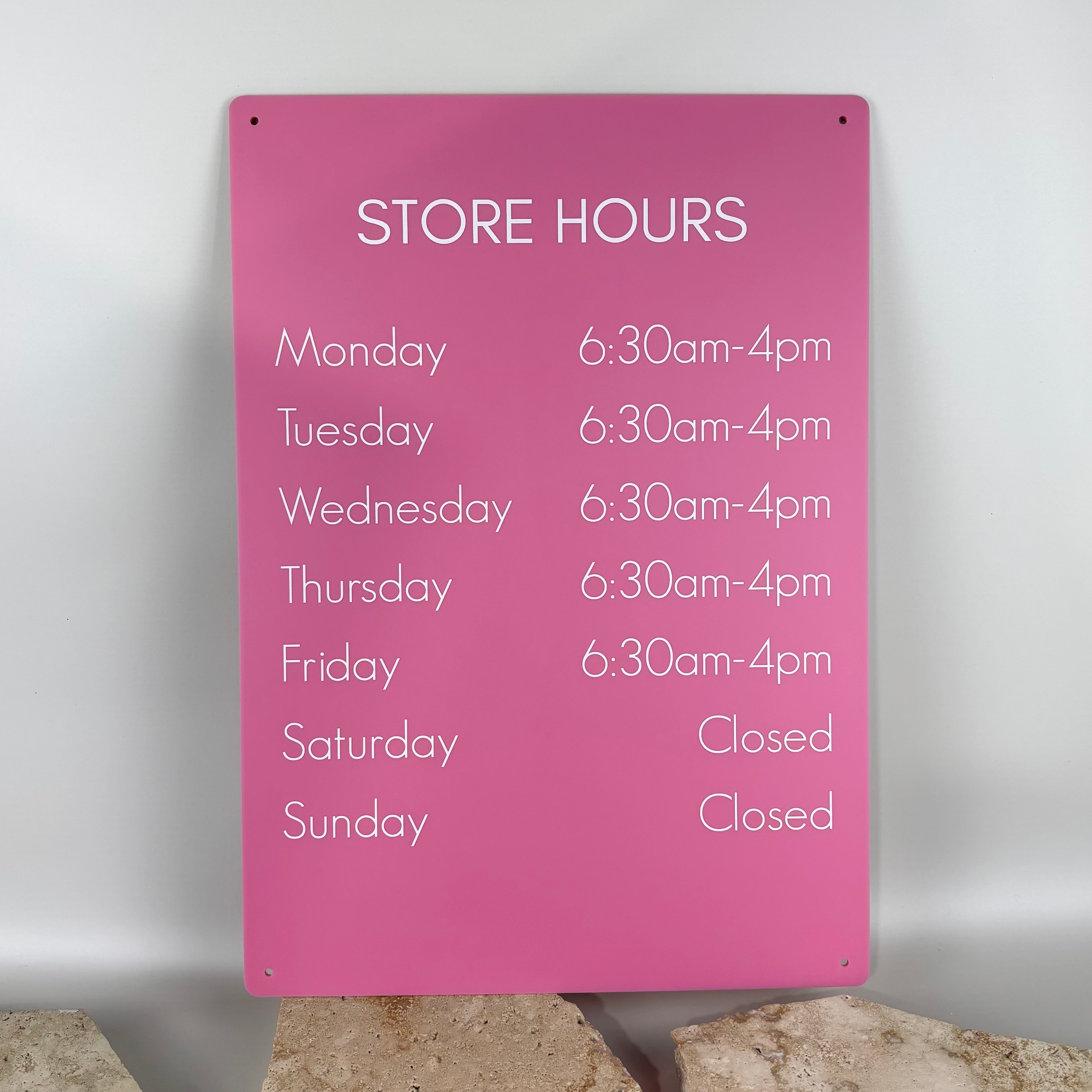 Bright pink acrylic store hours sign with white text listing opening times from Monday to Friday, 6:30am–4pm, and showing Saturday and Sunday as closed, displayed against a neutral background on stone slabs.