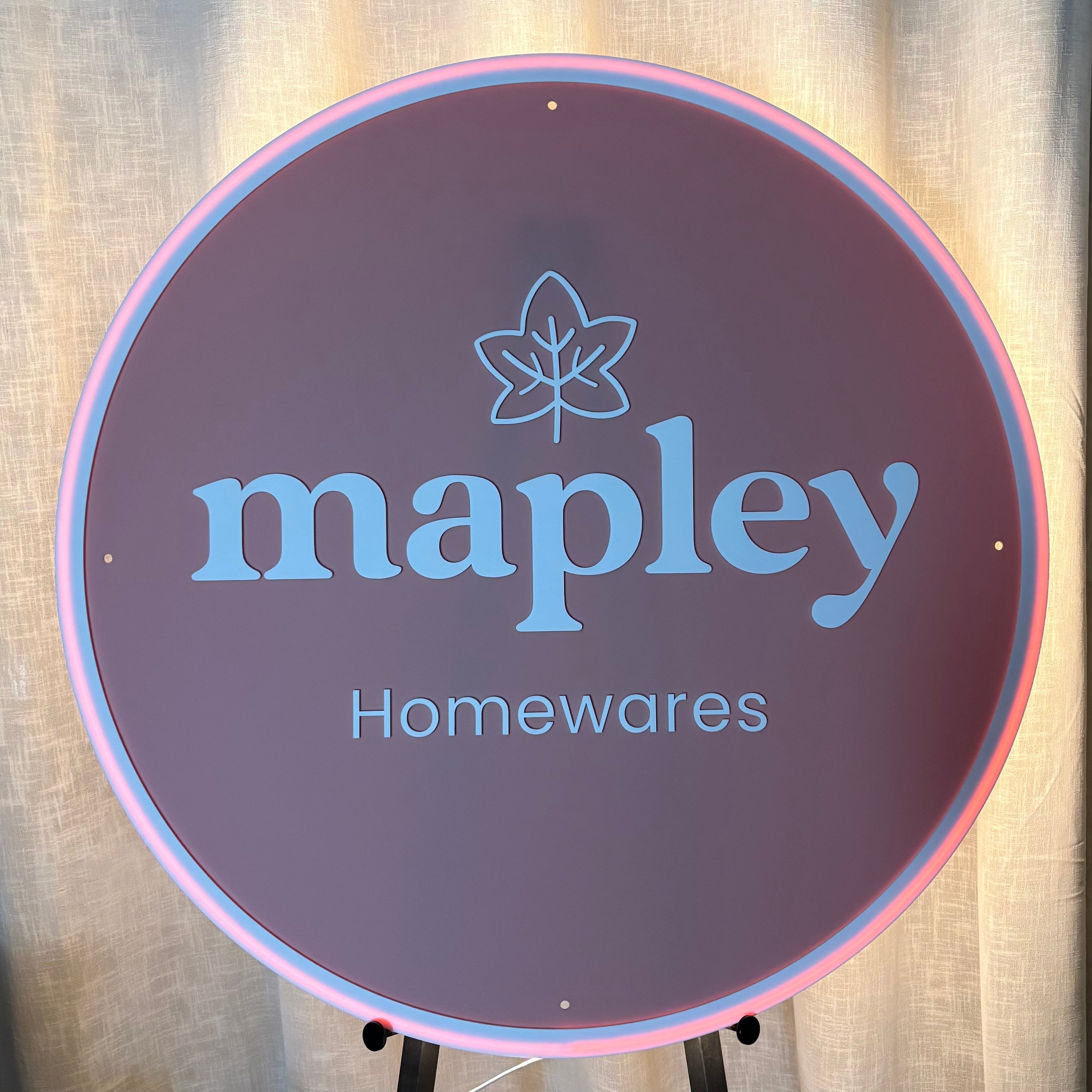 Large round backlit acrylic business sign featuring the Mapley Homewares logo with blue 3D acrylic lettering on a dark matte acrylic background, illuminated by a soft pink LED halo for a modern retail storefront display.