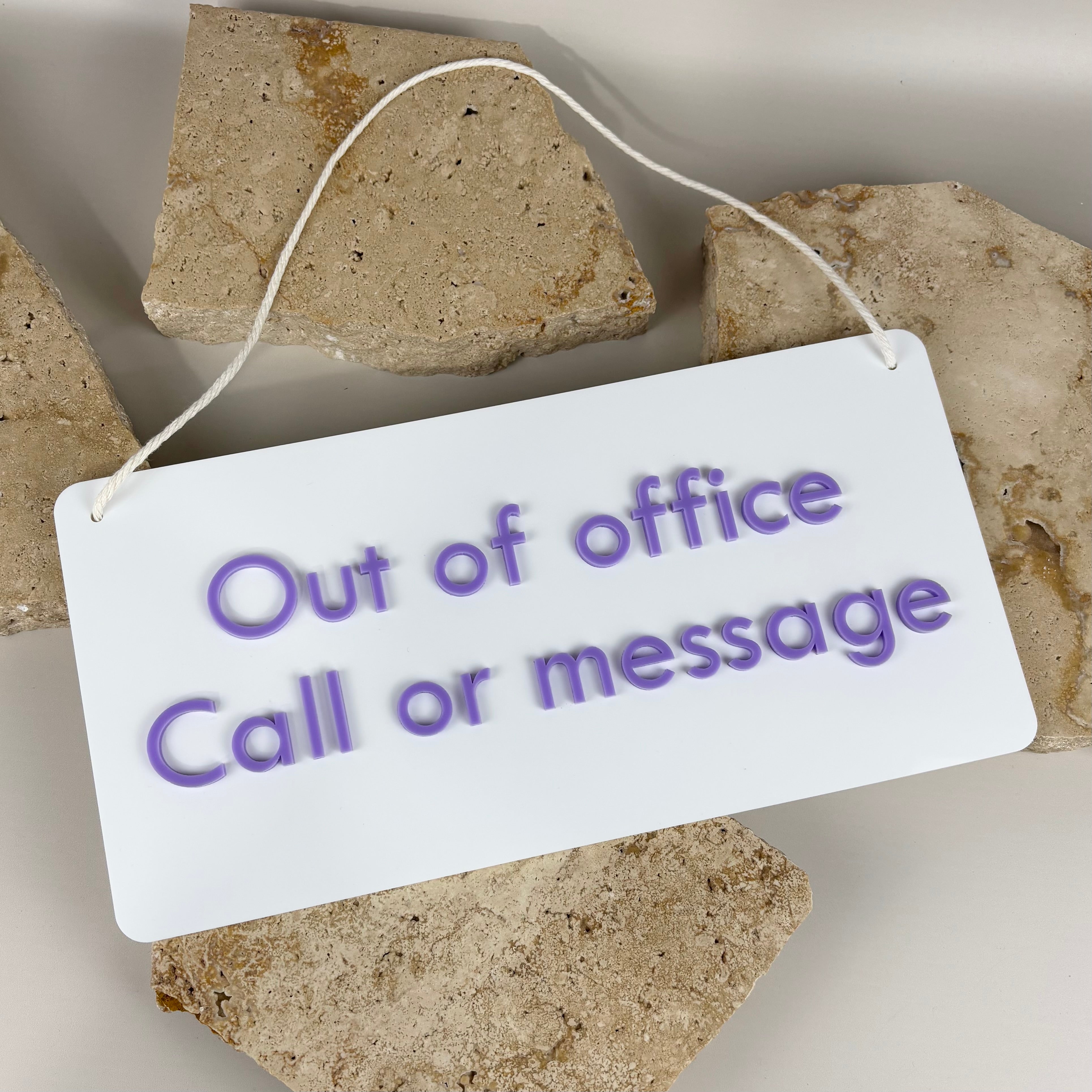 White acrylic hanging sign with rounded corners and lilac raised lettering that reads “Out of office Call or message,” displayed on natural stone slabs.