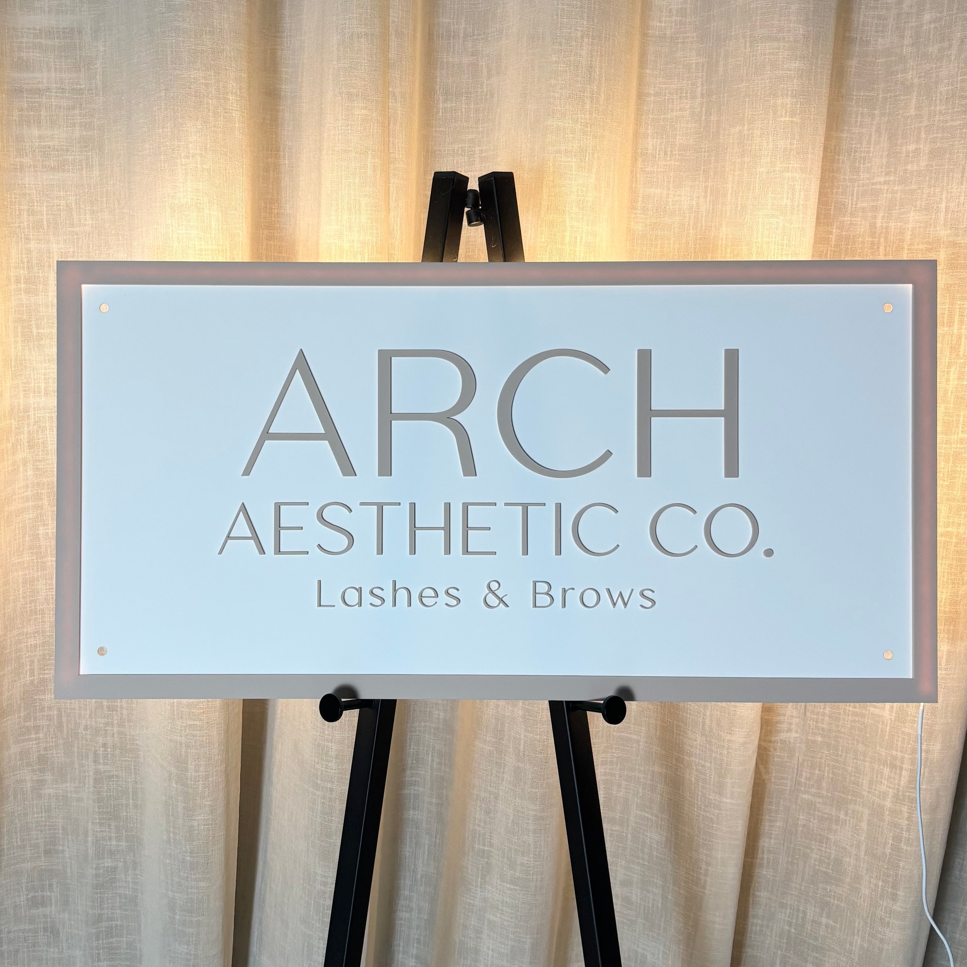 Backlit acrylic business sign with soft LED glow reading “Arch Aesthetic Co. Lashes & Brows,” mounted on an easel stand and styled against a neutral curtain backdrop