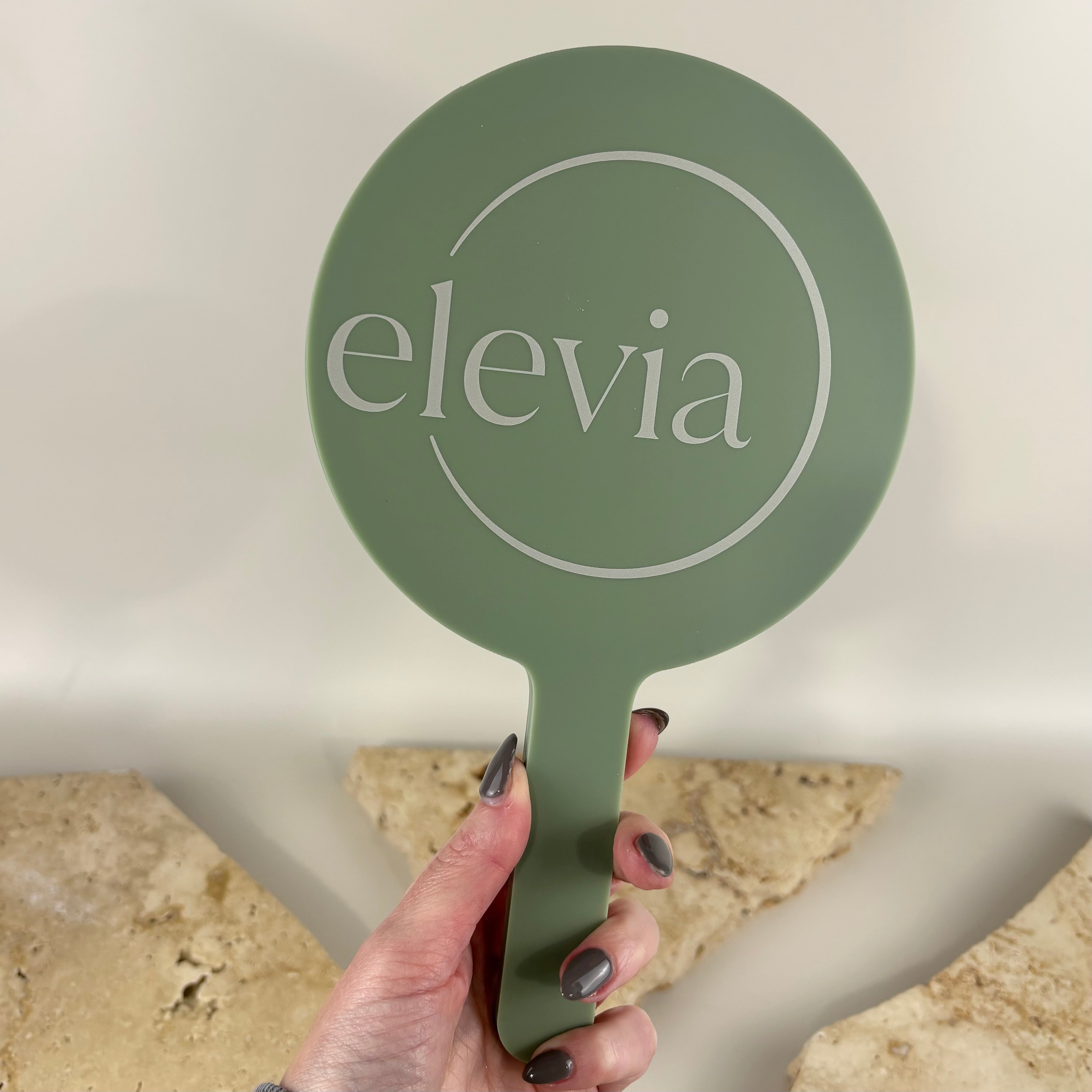 Custom sage green handheld salon mirror with UV printed white logo for Elevia. Premium acrylic hand mirror for beauty salons, hairdressers, cosmetic clinics and makeup artists, ideal for client consultations and showcasing finished treatments.