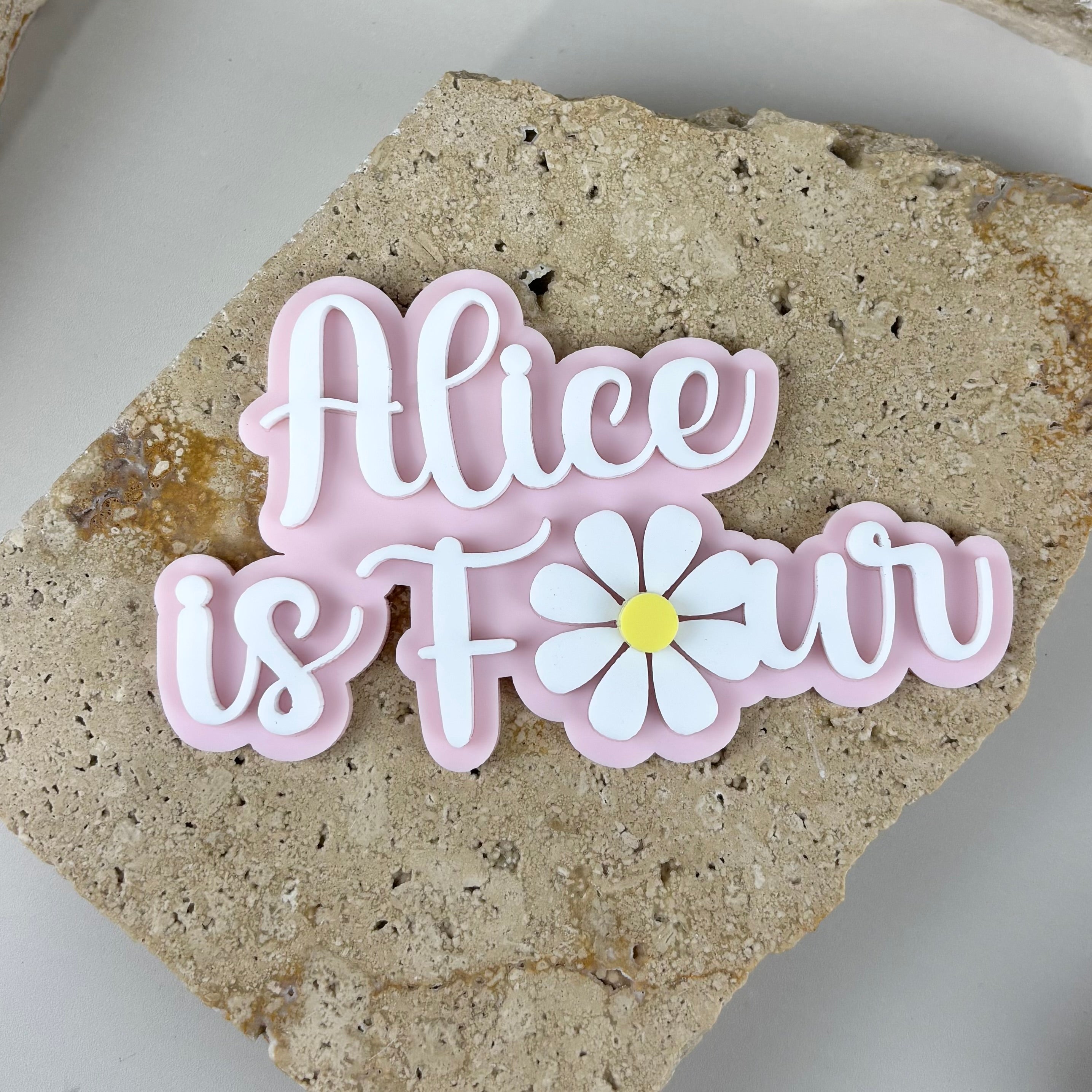 Custom acrylic cake topper with layered 3D lettering “Alice is Four”, personalised birthday cake fropper with pastel pink backing and white script, floral daisy design cake decoration for kids birthday celebration