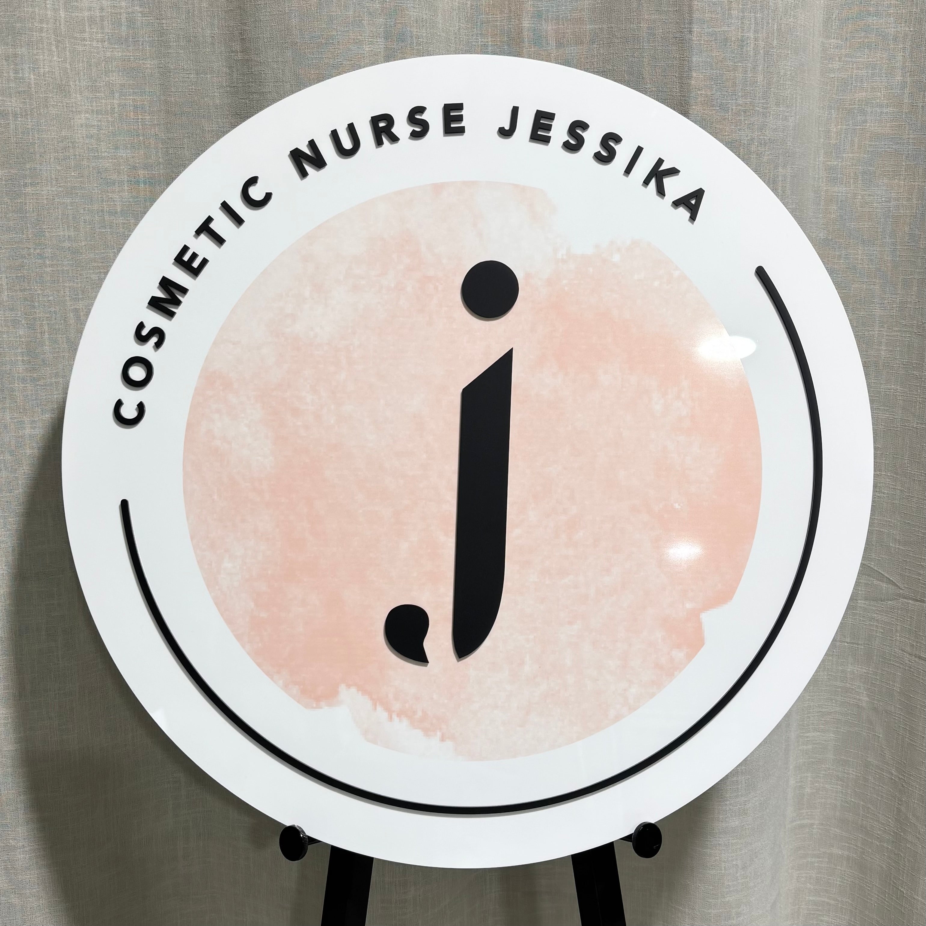 Custom round white acrylic business sign for Cosmetic Nurse Jessika with blush pink watercolour background and bold black initial logo, ideal for beauty clinics and professional branding