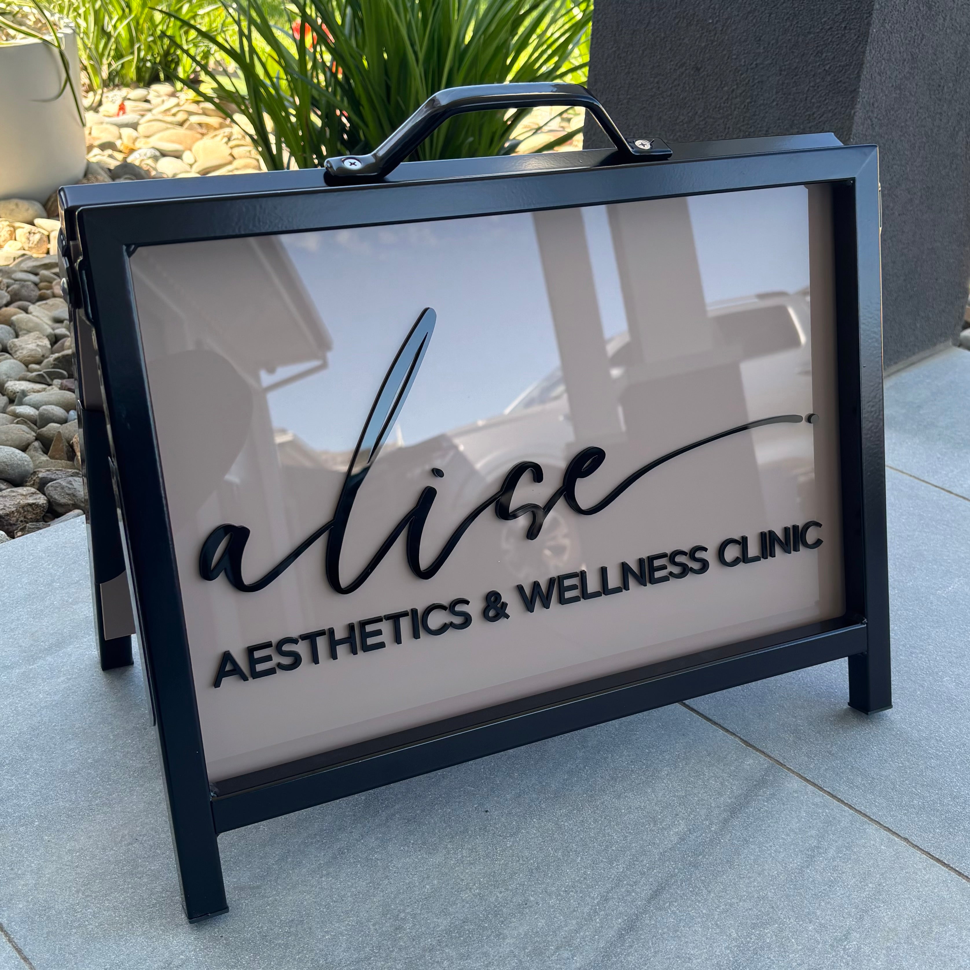 Custom black framed acrylic A-frame footpath sign with 3D lettering for “Alice Aesthetics & Wellness Clinic,” portable outdoor business signage displayed on concrete with garden backdrop.