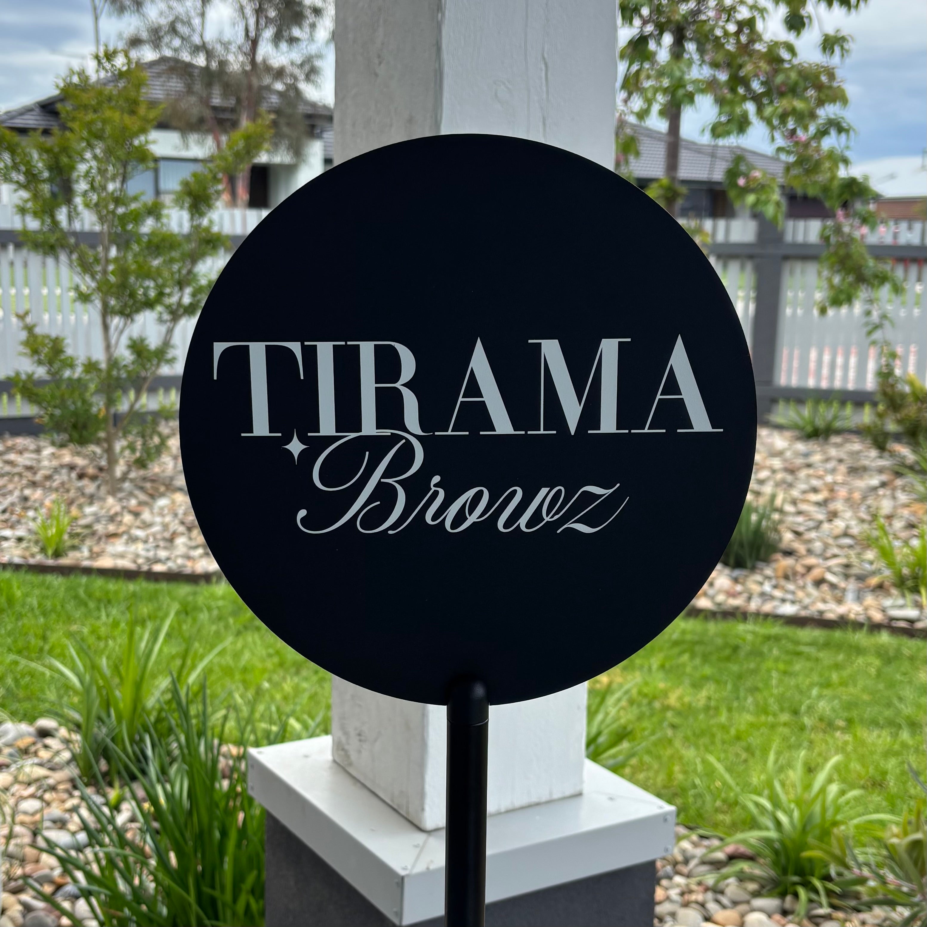 Close up of a black round acrylic business sign with white logo text reading “Tirama Browz”, mounted on a freestanding pole in an outdoor garden setting.