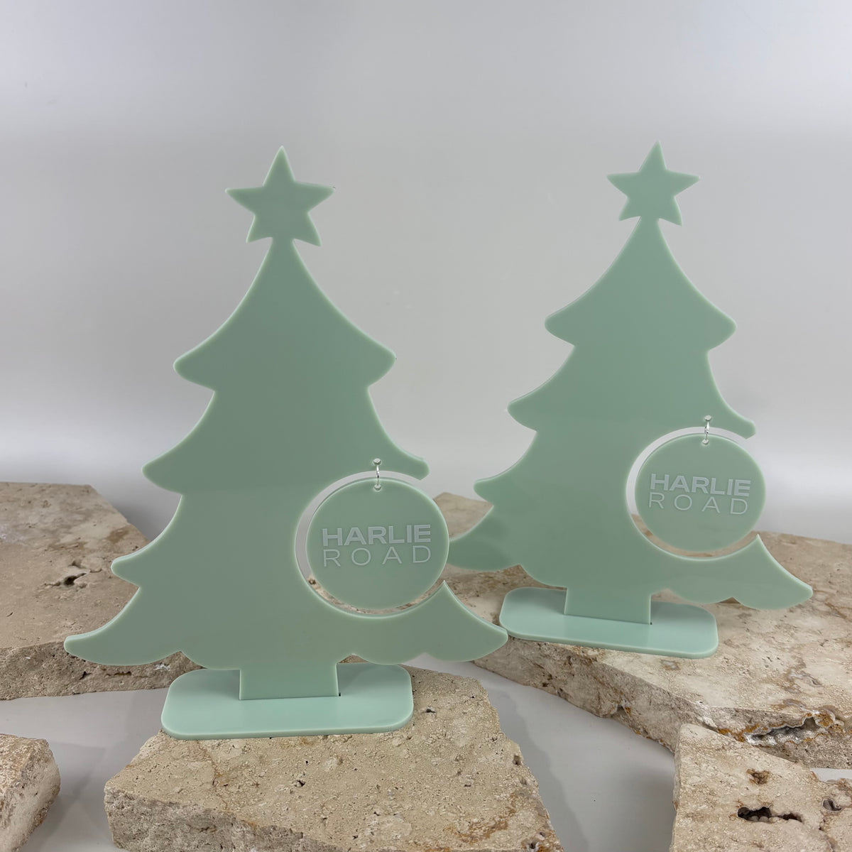 Two custom sage green acrylic Christmas tree display stands with star toppers, freestanding bases and personalised hanging logo ornaments for Harlie Road.