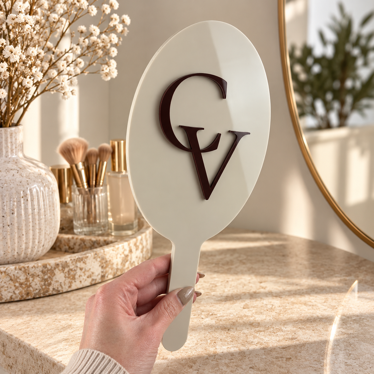 Custom cream oval handheld mirror with raised dark brown GV logo, styled in a bright modern beauty studio with warm natural lighting.
