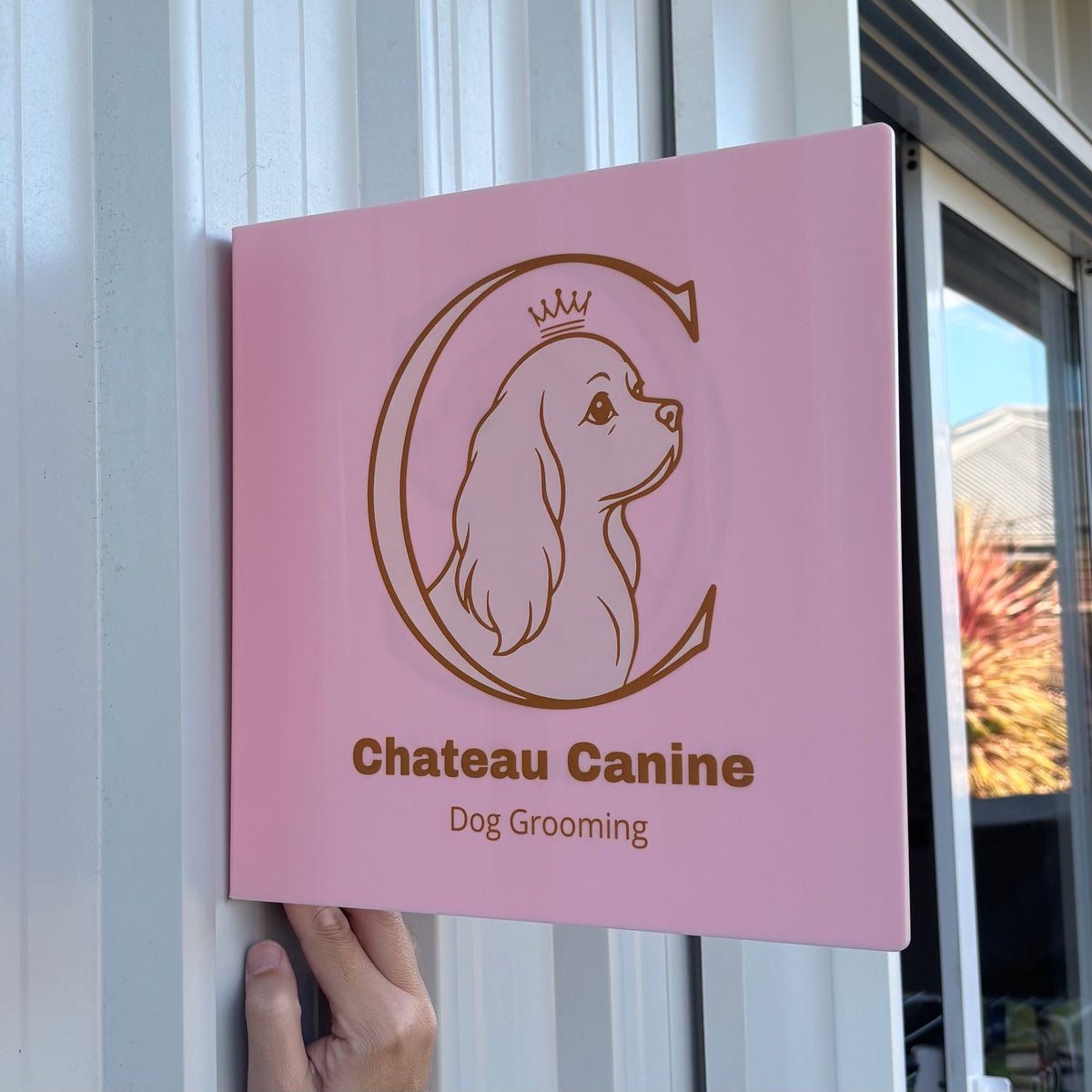 Custom square pink acrylic business sign featuring the Chateau Canine dog grooming logo and gold printed lettering, displayed outside a salon entrance.