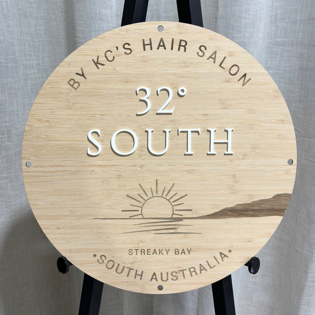 Custom round bamboo business sign for 32° South hair salon, featuring laser engraved branding, a sunset design and raised white acrylic lettering.