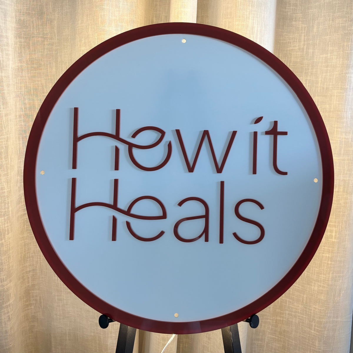 Custom round LED backlit acrylic business sign for How It Heals, featuring a white illuminated face, burgundy 3D logo lettering and matching burgundy circular border.