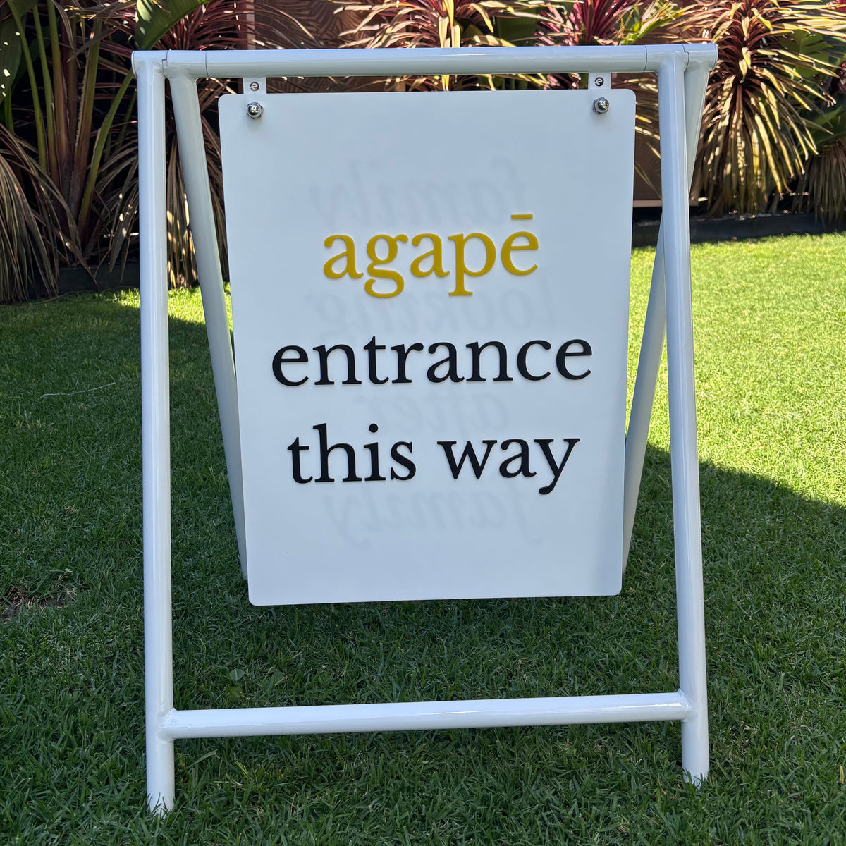Custom white swing pavement sign for Agapē featuring raised yellow and black acrylic lettering reading entrance this way, displayed outdoors in a white metal frame.