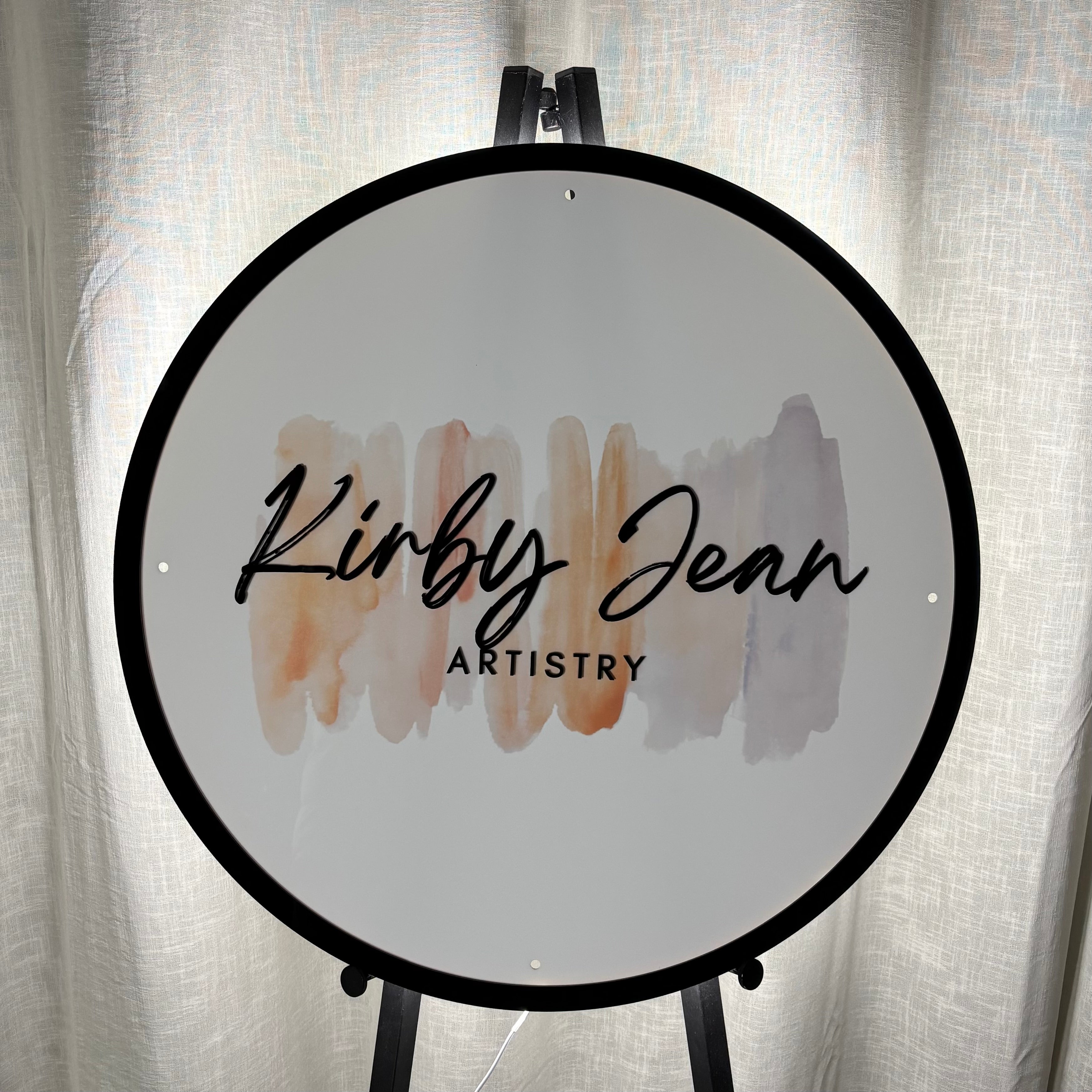 Custom round acrylic business sign for “Kirby Jean Artistry,” featuring black script lettering over a soft neutral watercolour background and a sleek black border, displayed on a stand — ideal for salons, makeup studios, or beauty businesses.