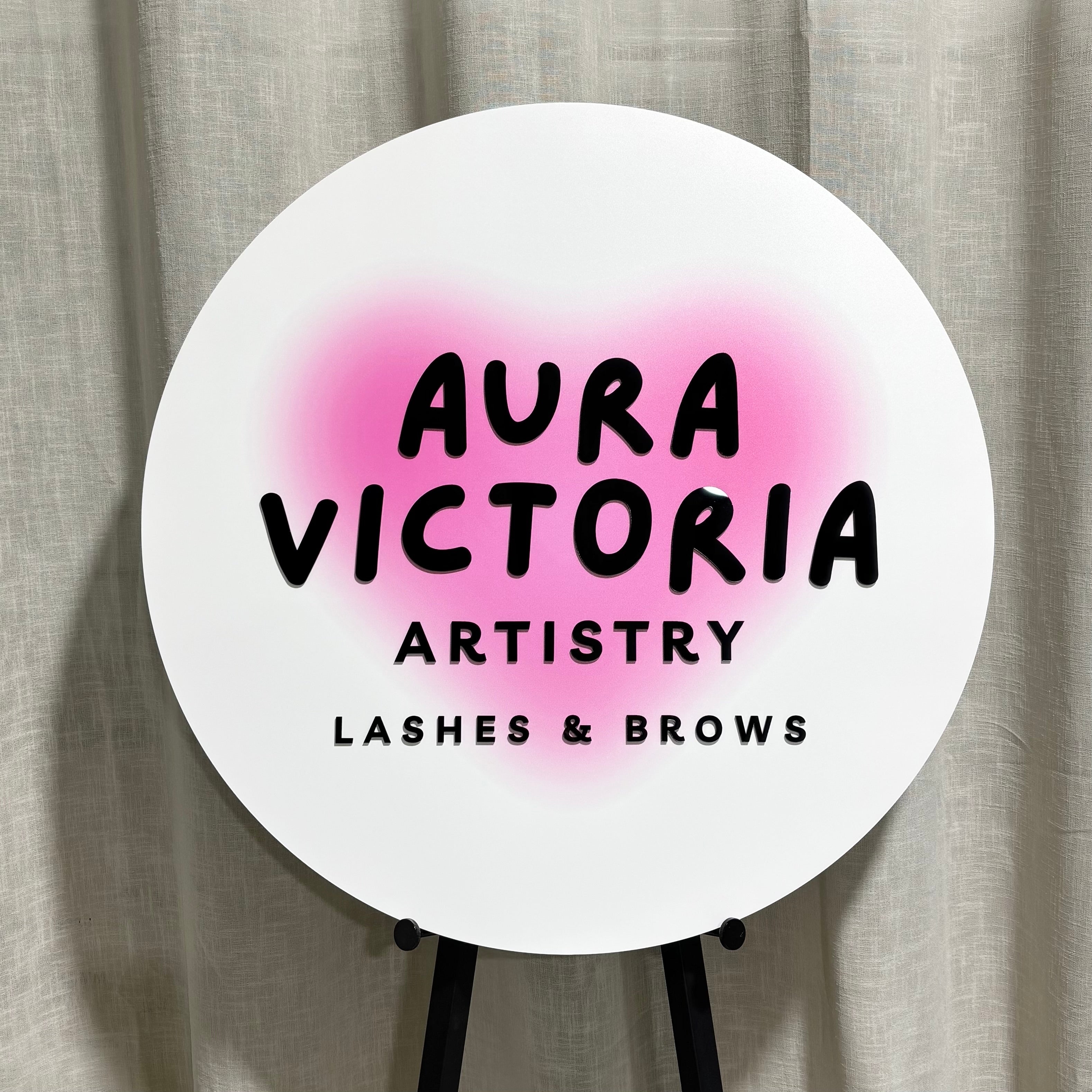Large round white acrylic business sign featuring black 3D acrylic lettering with a pink UV printed background, custom made for a lash and brow salon to create a modern, eye-catching storefront or reception display.