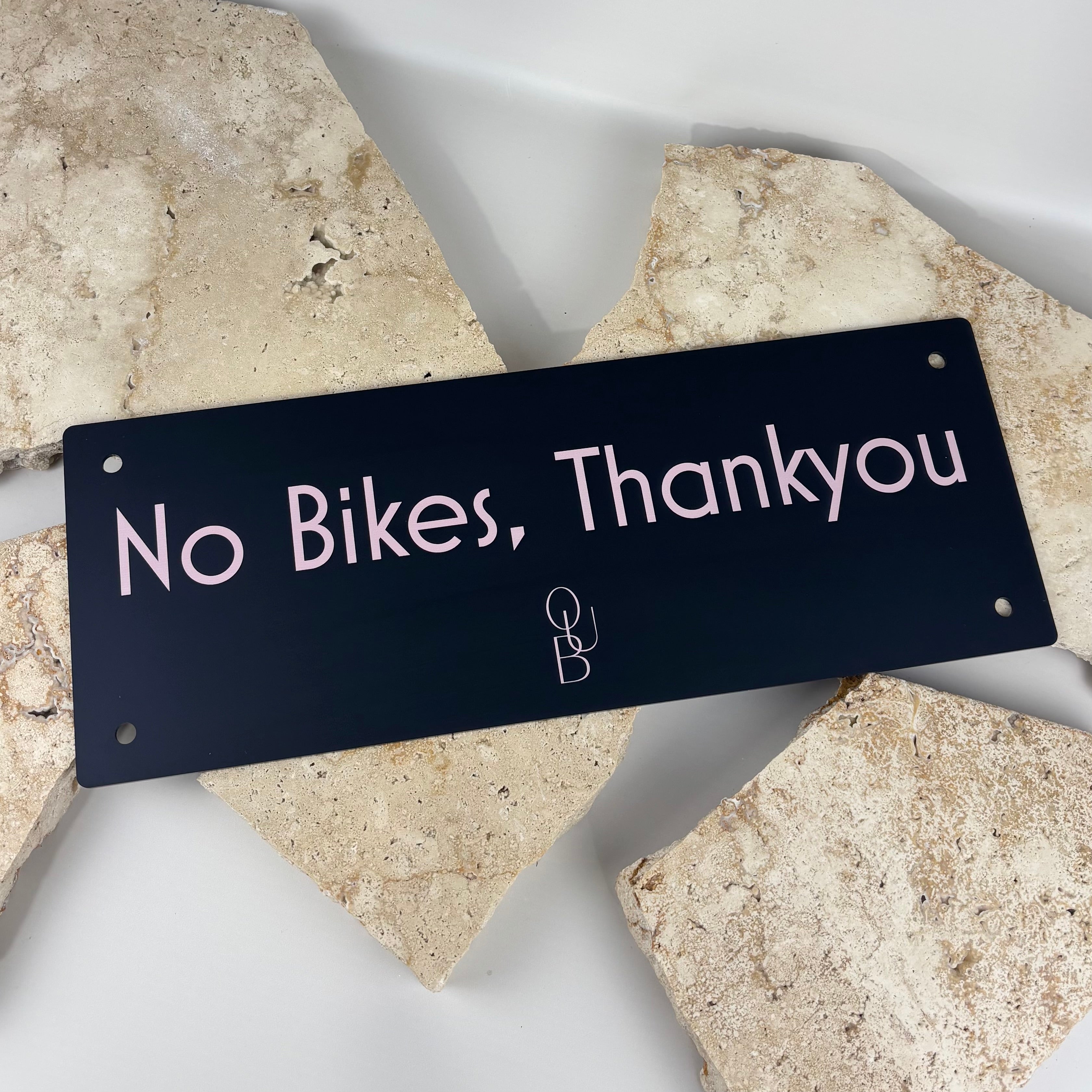 Black acrylic rectangular sign reading “No Bikes, Thankyou” with soft pink lettering and rounded corners, featuring pre-drilled mounting holes, styled on a neutral stone background
