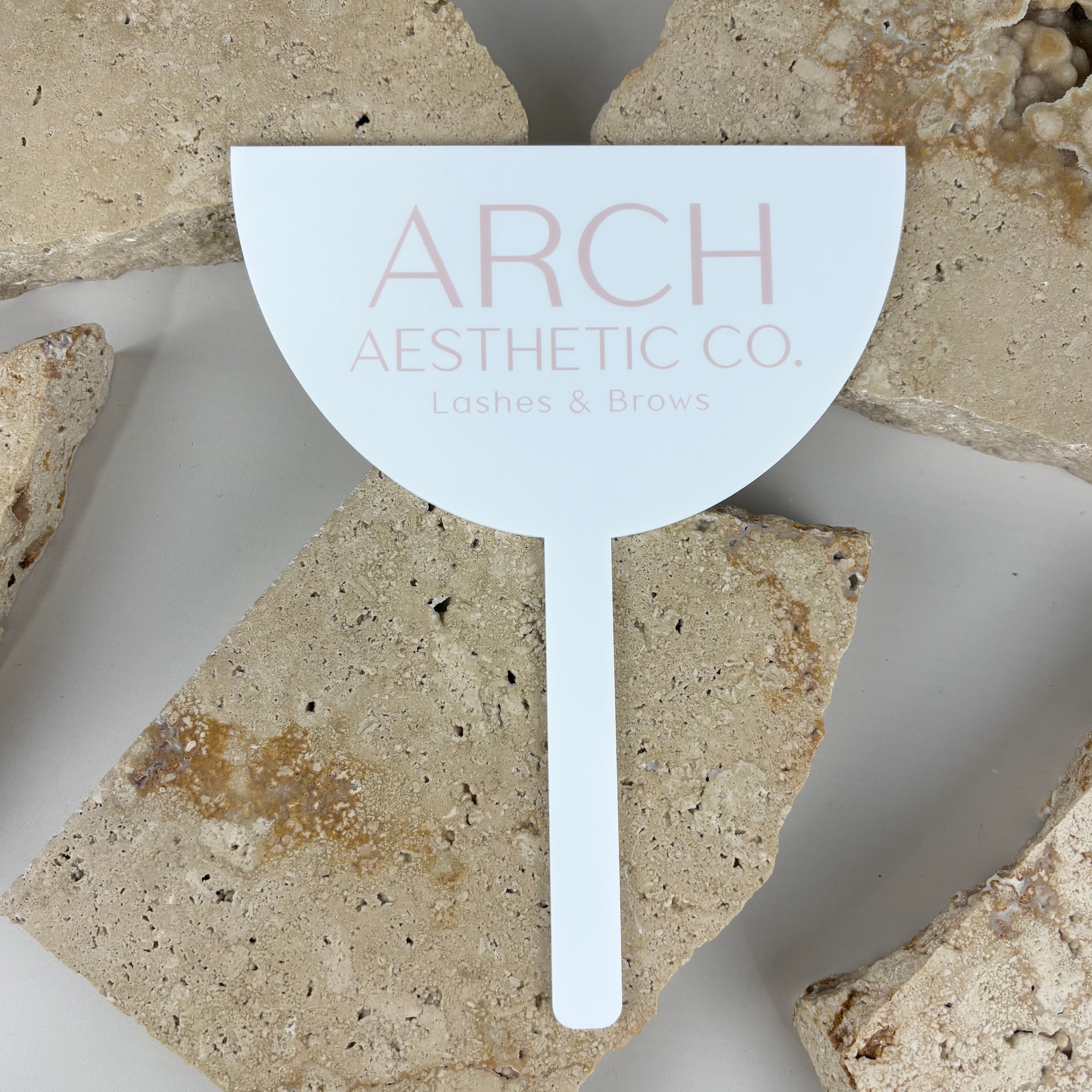 Frosted acrylic face shield with arch shape and soft pink logo reading Arch Aesthetic Co Lashes & Brows, styled on neutral stone background for salon photography use