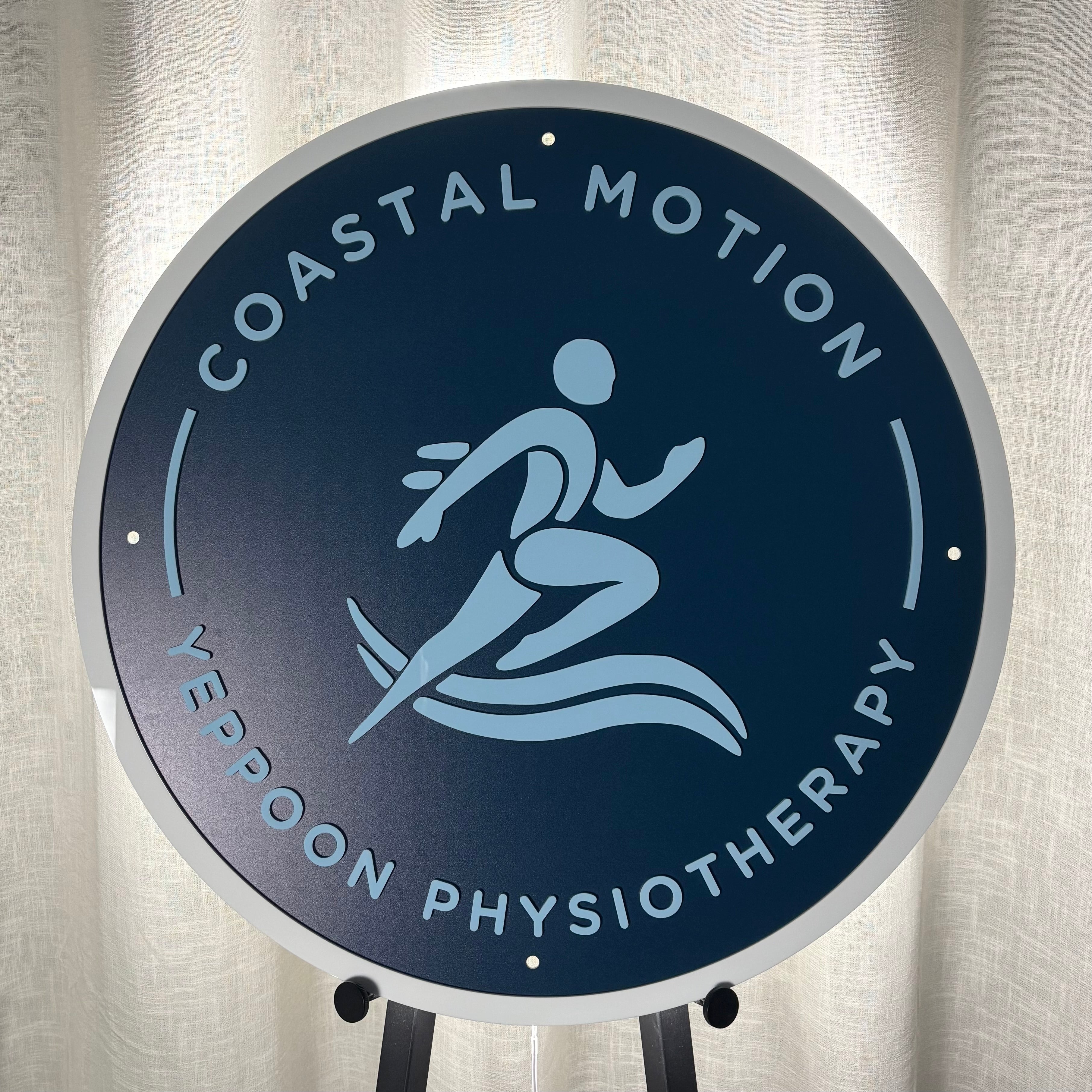 Round acrylic business sign for Coastal Motion Yeppoon Physiotherapy, featuring UV printed graphics in blue tones on a matte navy backing, displayed on an easel for clinic branding.