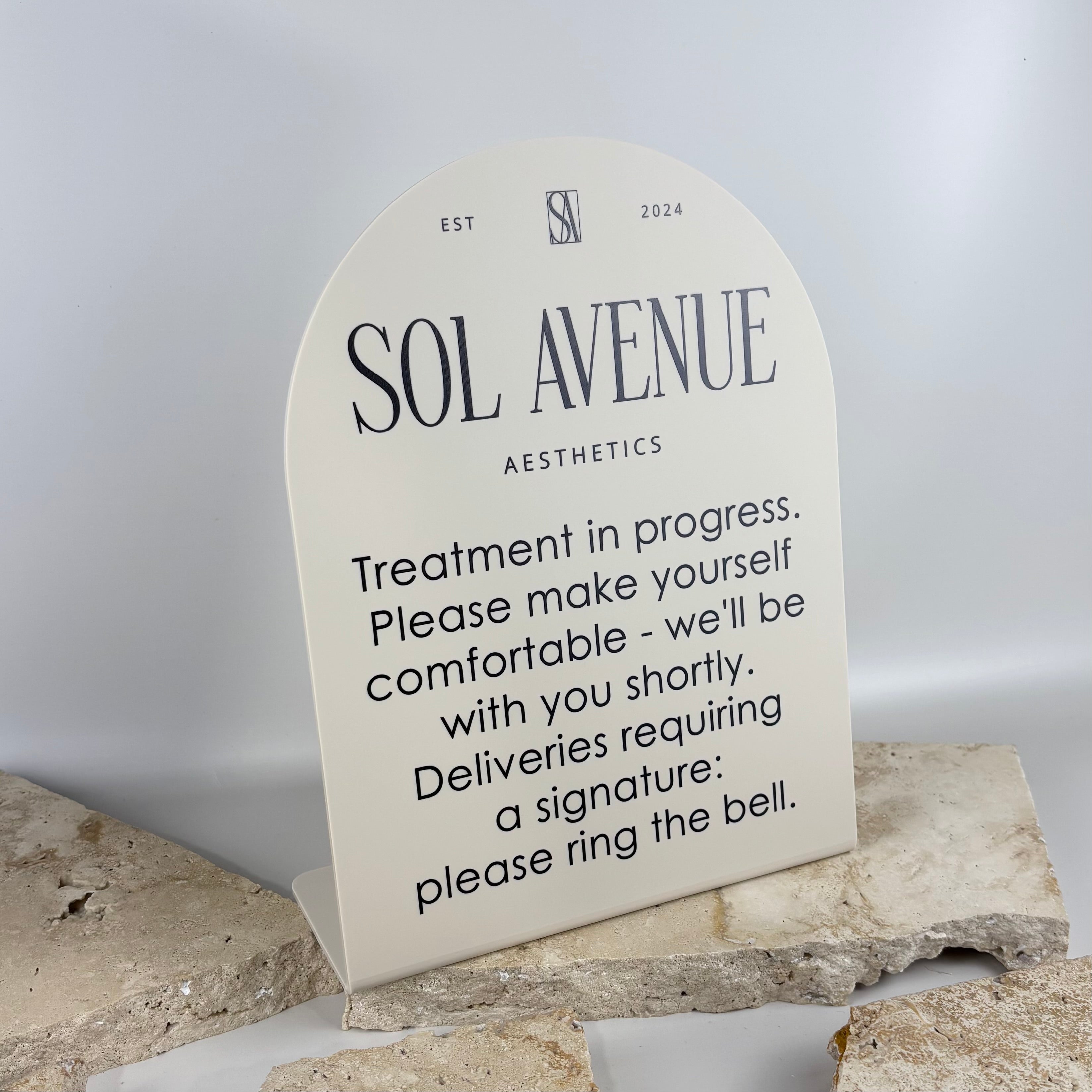 “Curved acrylic desk sign for beauty salon reception, custom UV printed business signage with ‘treatment in progress’ message, aesthetic clinic counter display, modern neutral arch stand for client communication and branding