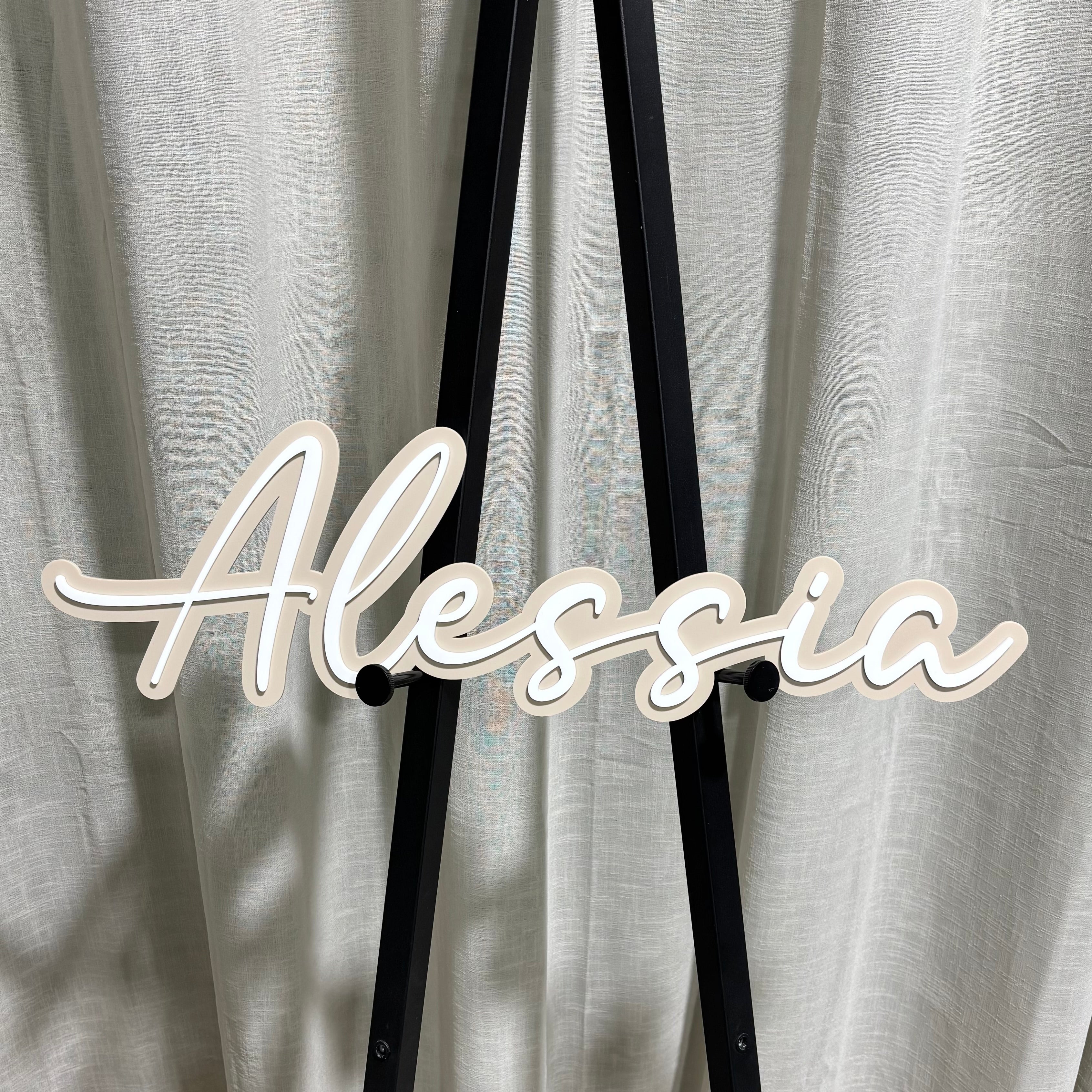 custom acrylic name sign in scripted font reading “Alessia” displayed on black stand, layered white and beige 3D lettering for birthdays events and celebrations australia