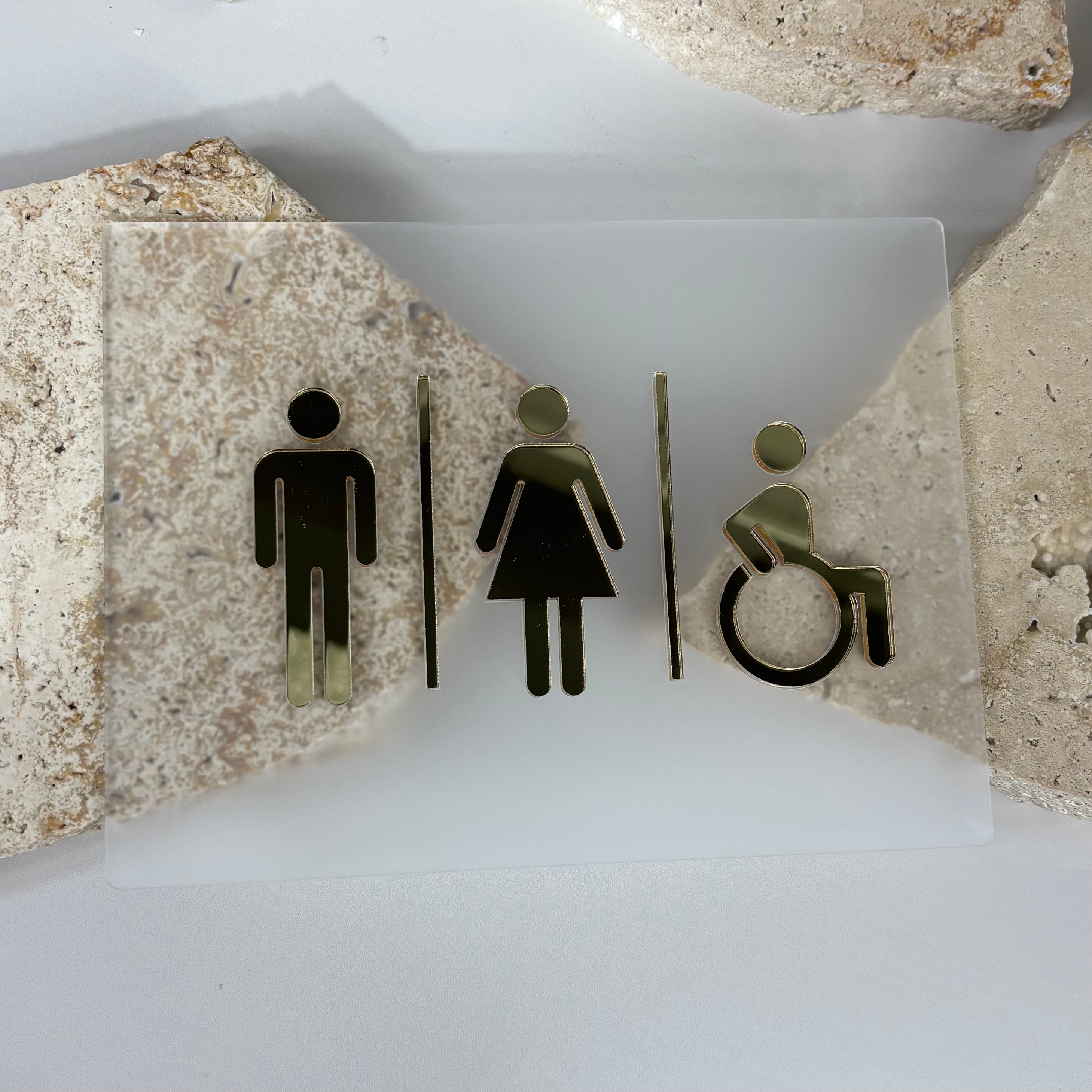 Unisex and accessible bathroom sign made from frosted acrylic with mirrored gold male, female, and wheelchair icons, displayed on neutral stone background.
