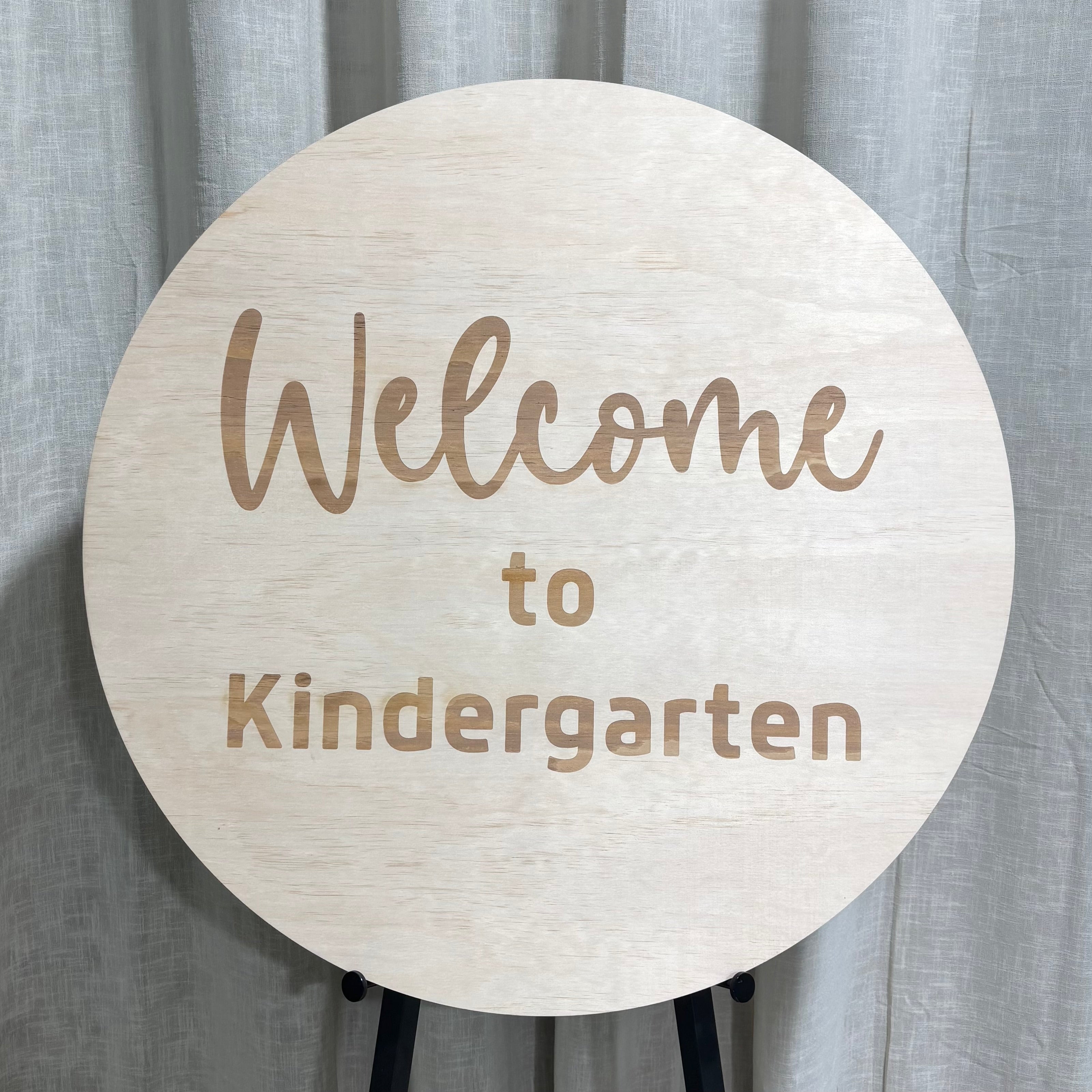 Round wooden welcome sign reading ‘Welcome to Kindergarten’ in engraved lettering, natural timber classroom sign displayed on an easel.