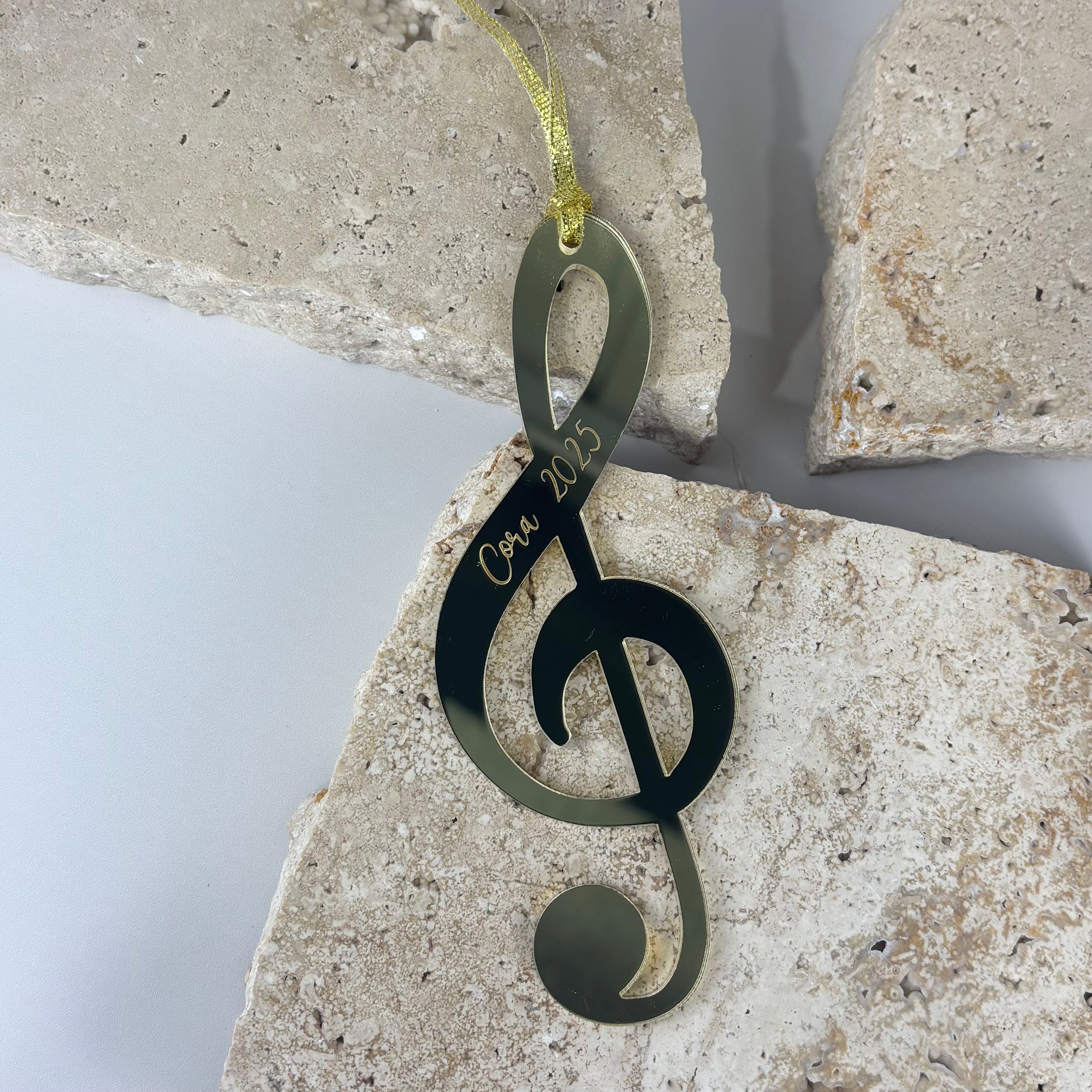 Gold mirrored acrylic treble clef ornament with “Coor 2025” engraving, displayed on textured stone tiles with a gold hanging ribbon in a clean studio setup.