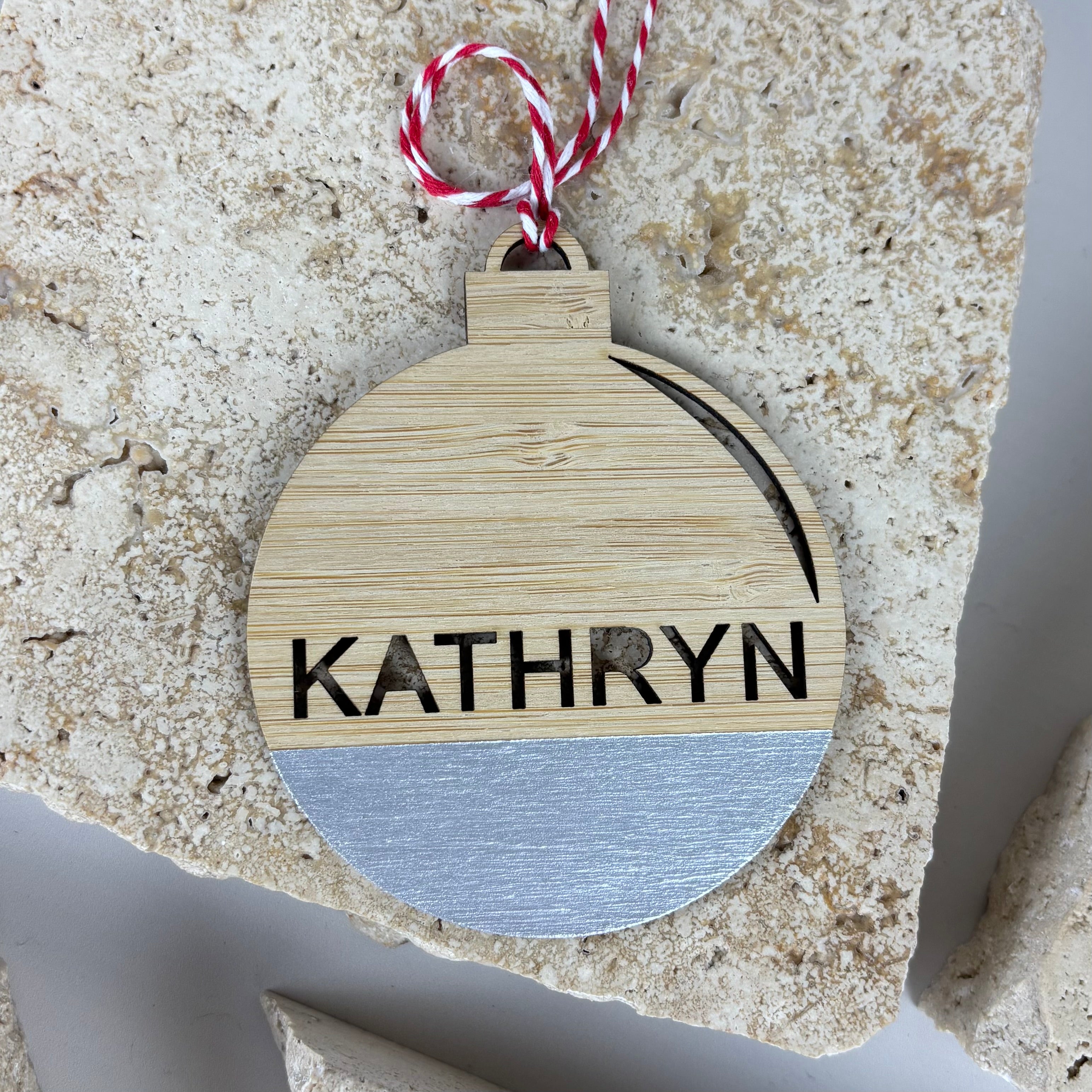 Personalised bamboo Christmas bauble engraved with the name “Kathryn”, natural wood ornament with silver painted base and red white hanging twine.