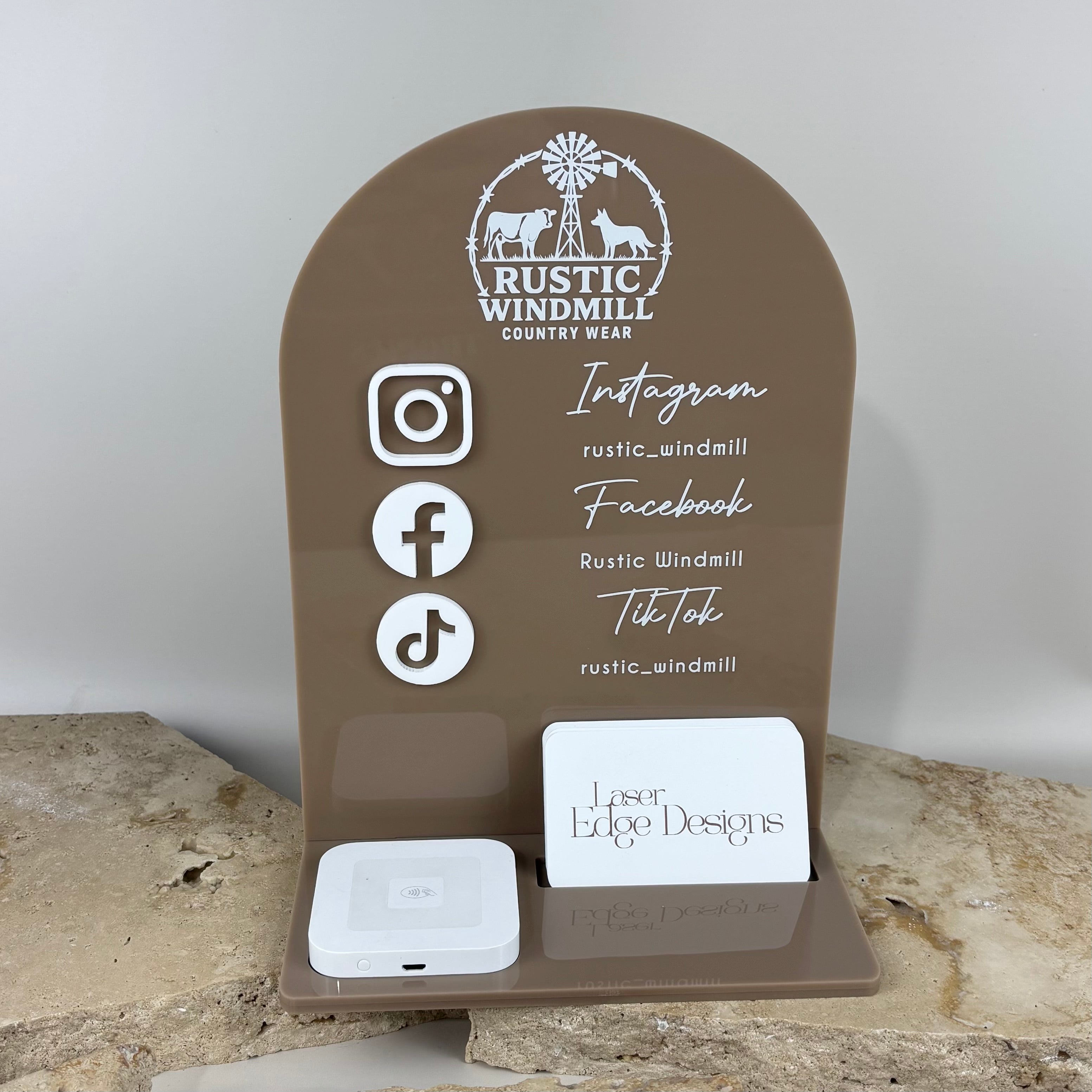 Brown acrylic arch business stand with raised social media icons, NFC tap device and business card holder, featuring Rustic Windmill Country Wear branding, displayed on a neutral stone background