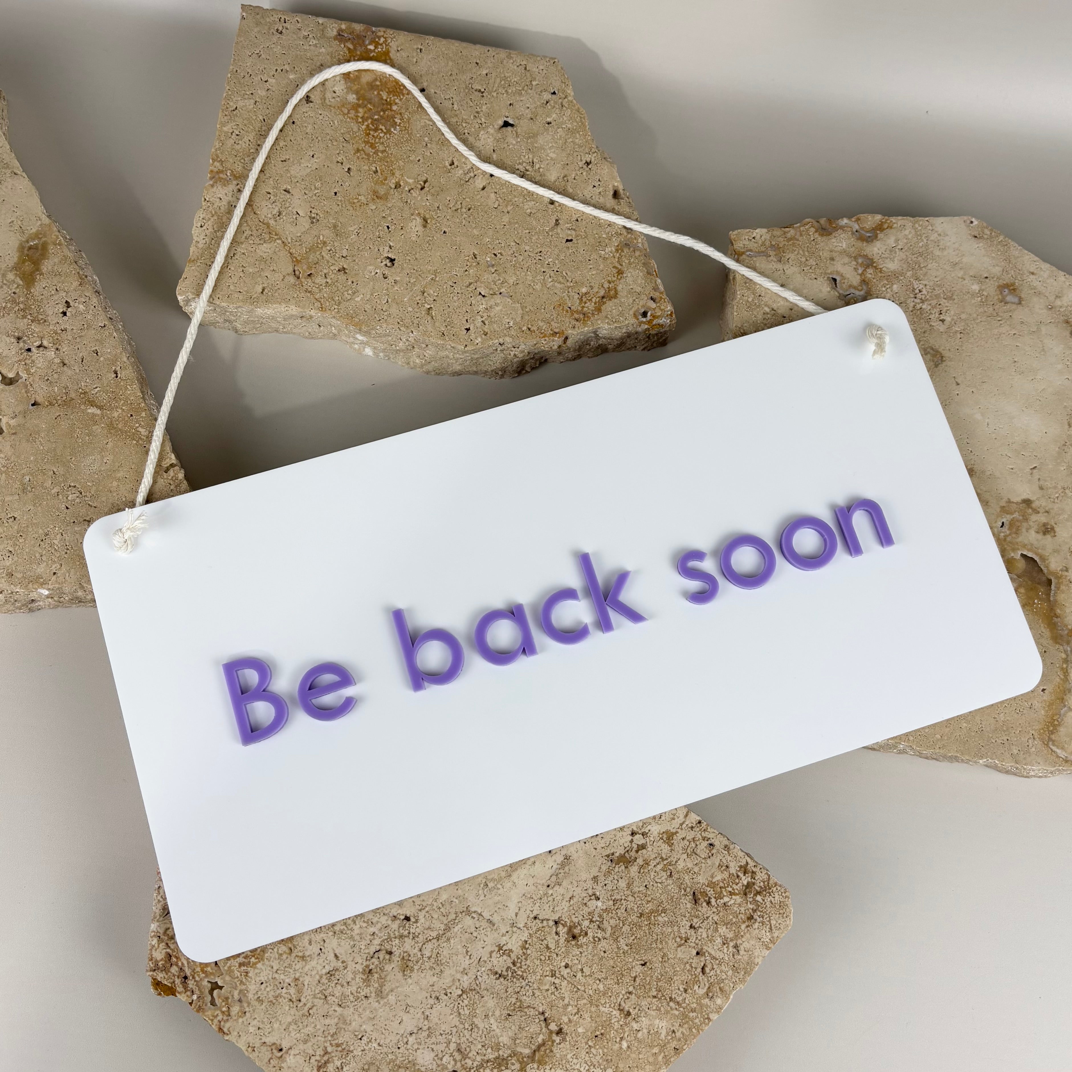 White acrylic hanging sign with lilac raised lettering that reads “Be back soon,” placed on neutral stone slabs.